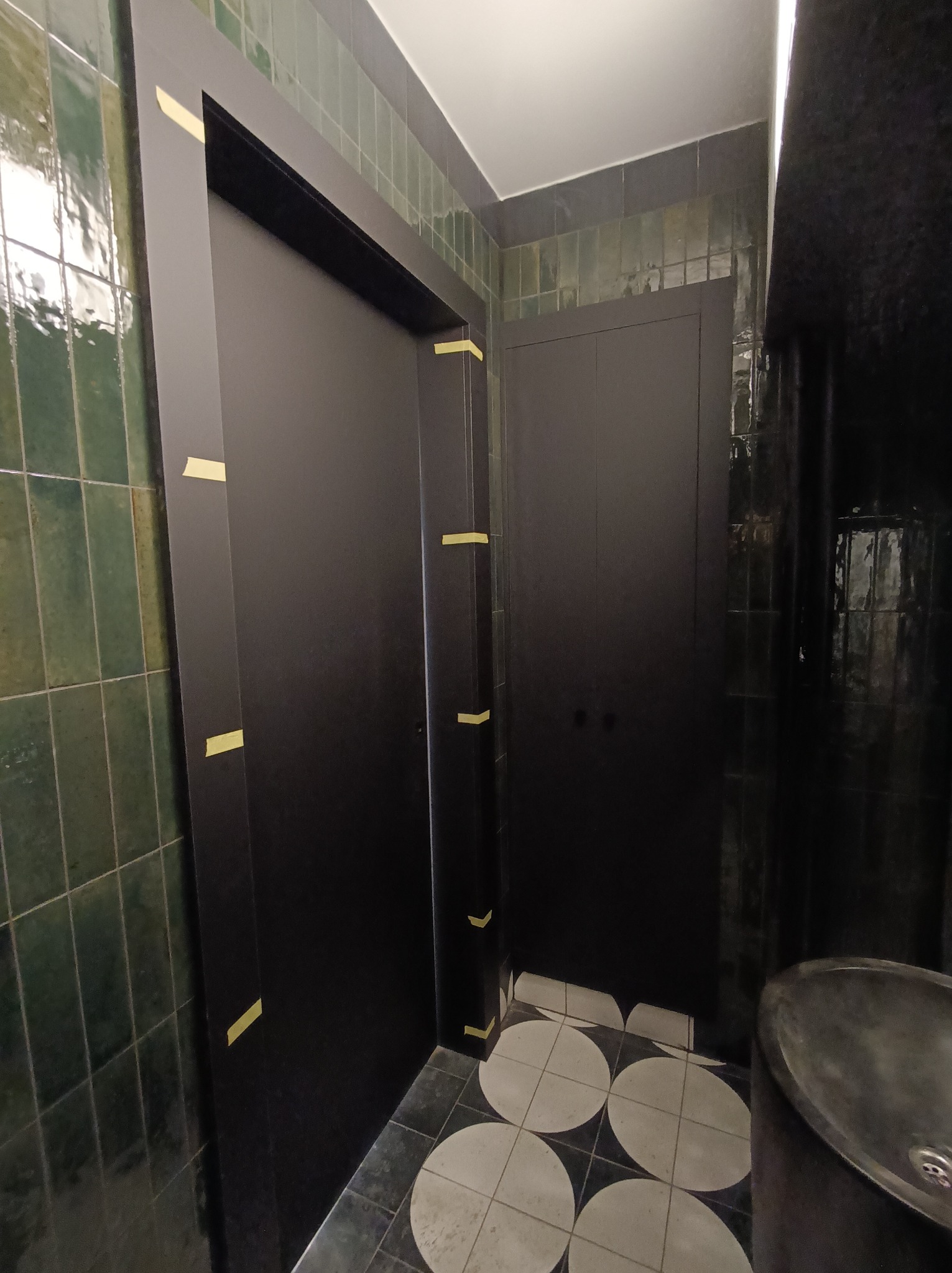 Czarna szafa wnękowa z taśmą malarską zabezpieczającą, zielone kafelki na ścianie, okrągłe płytki na podłodze i umywalka z szarego kamienia.