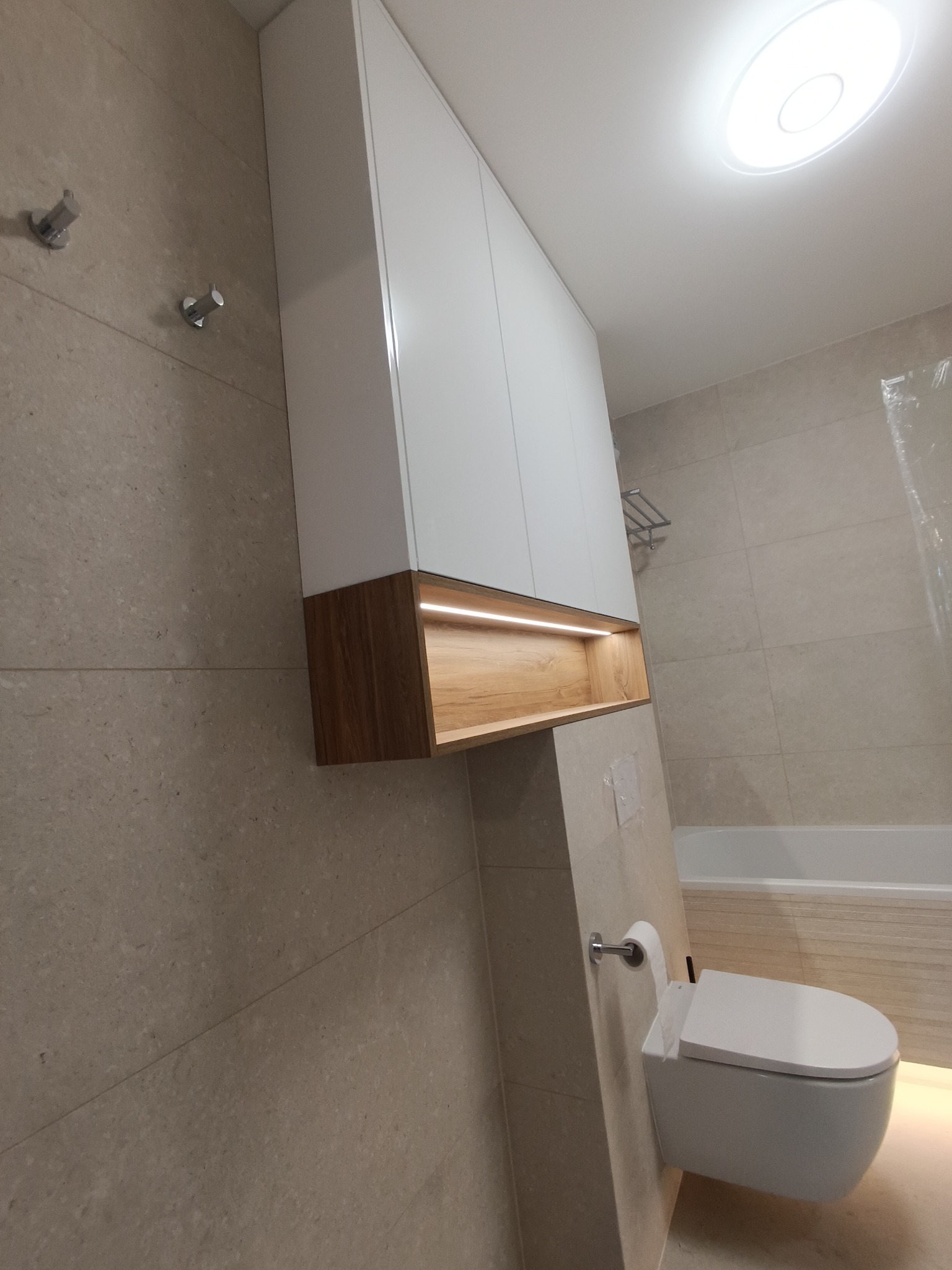 Biała szafka łazienkowa z drewnianym akcentem i podświetleniem LED nad toaletą podwieszaną w nowoczesnej łazience wykończonej beżowymi płytkami.