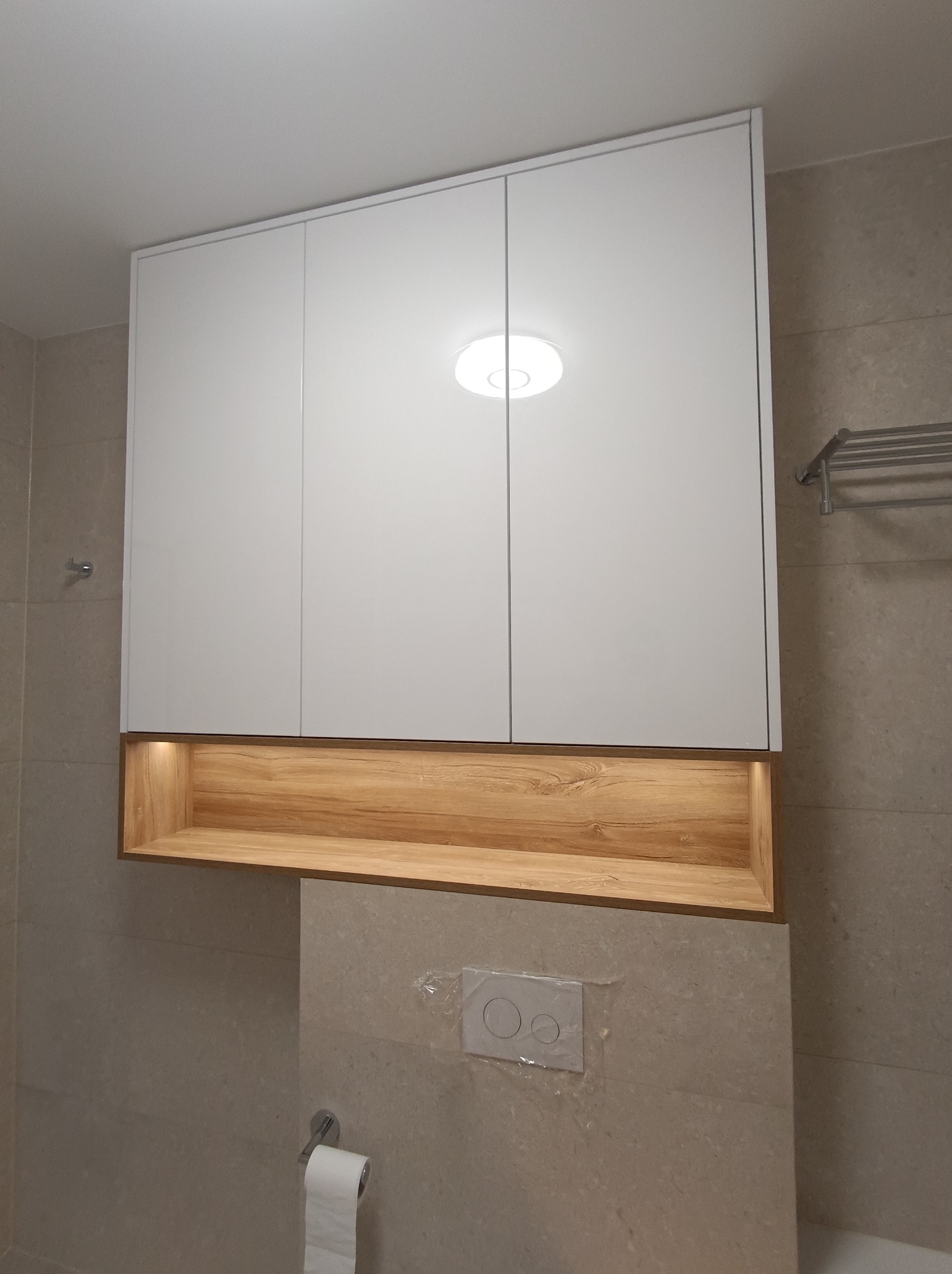 Biała szafka łazienkowa z trzema drzwiczkami i drewnianą wnęką z oświetleniem, zamontowana nad toaletą w łazience wyłożonej beżowymi płytkami.