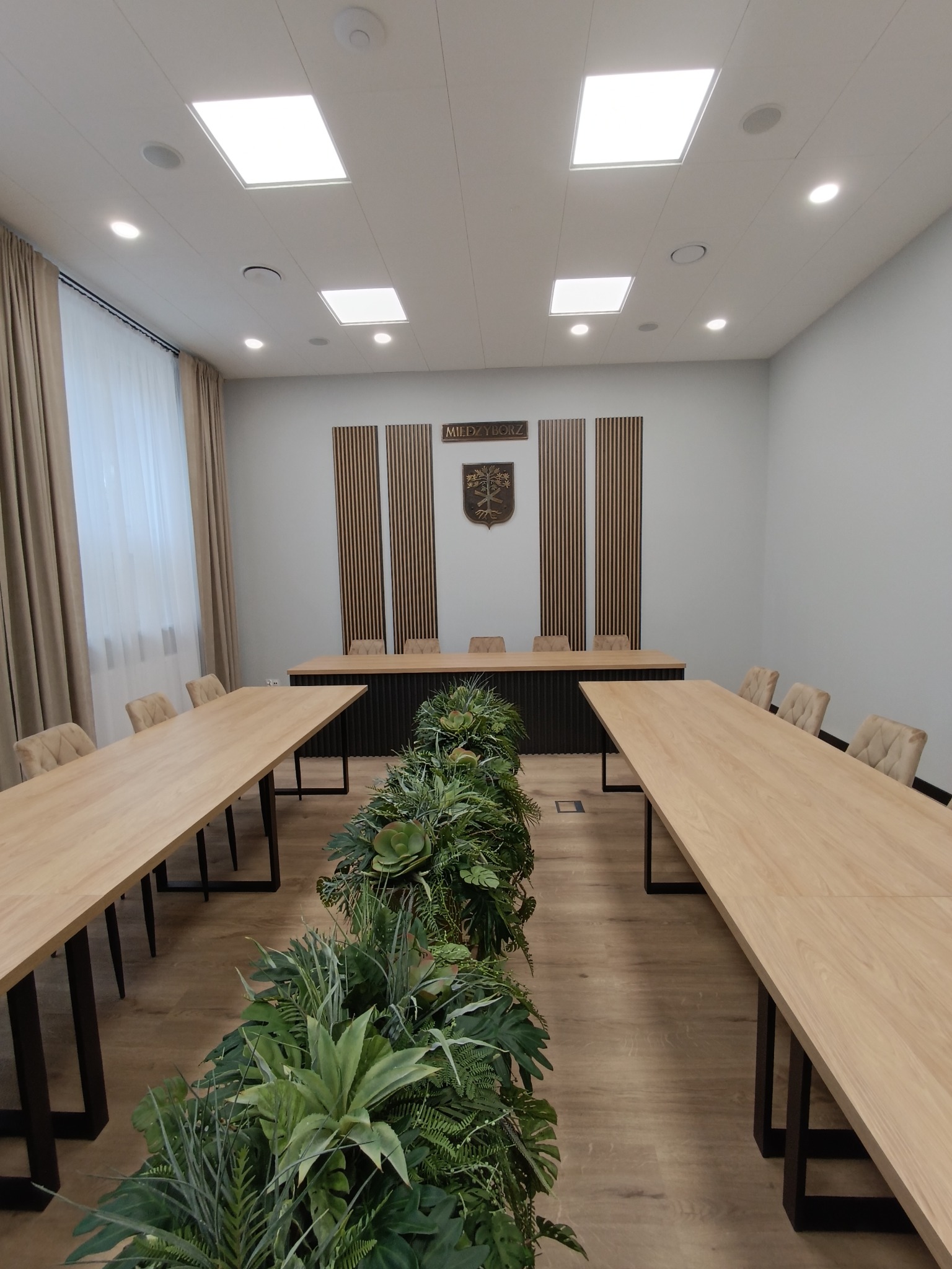Sala konferencyjna z długimi, jasnymi stołami z czarnymi nogami, krzesłami z beżową tapicerką, centralną dekoracją z zielonych roślin oraz herbem miasta na ścianie.