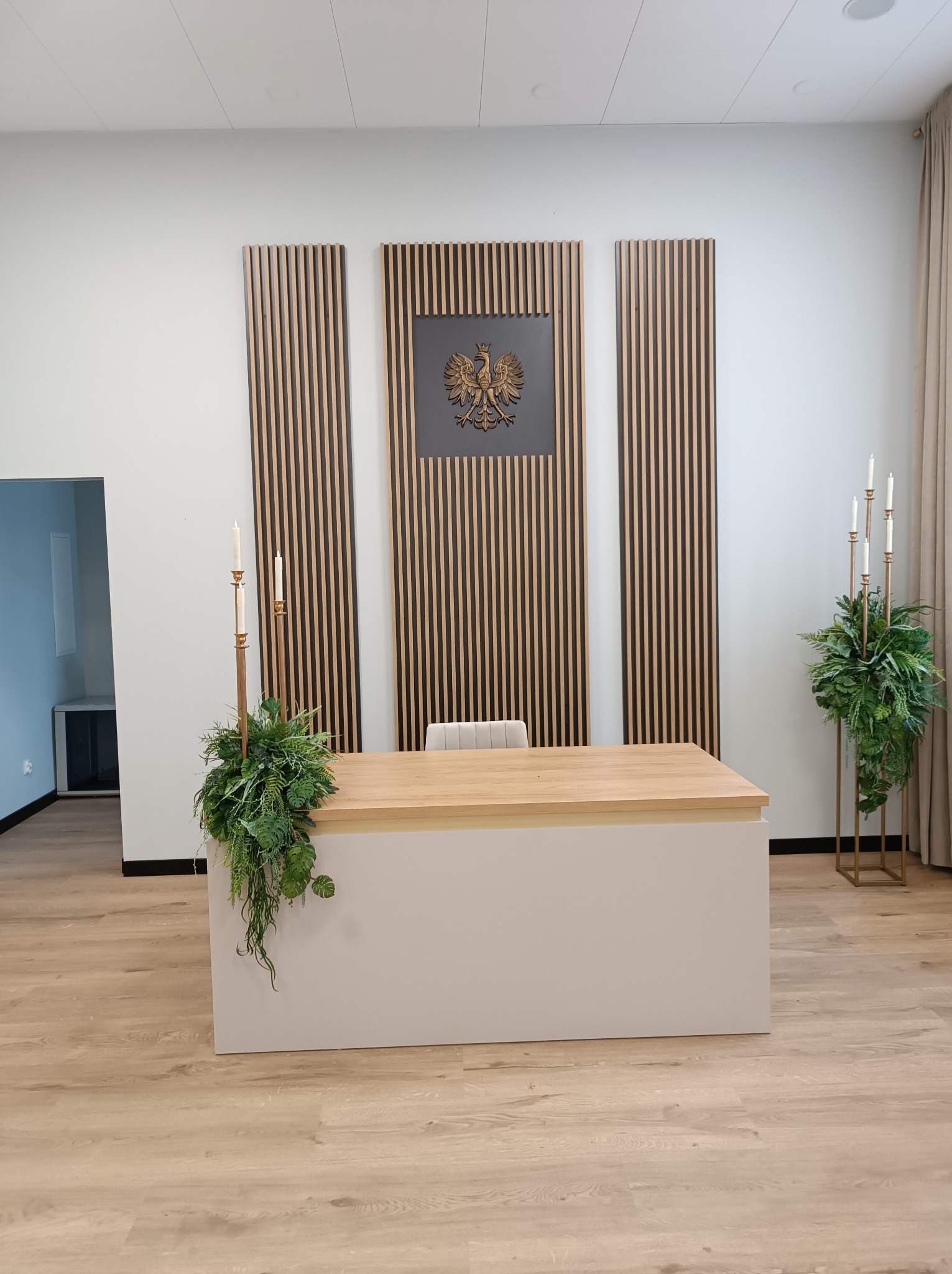 Stół recepcyjny z drewnianym blatem i białym frontem, dekorowany roślinami i świecami, w tle ściana z drewnianymi panelami i godłem Polski.