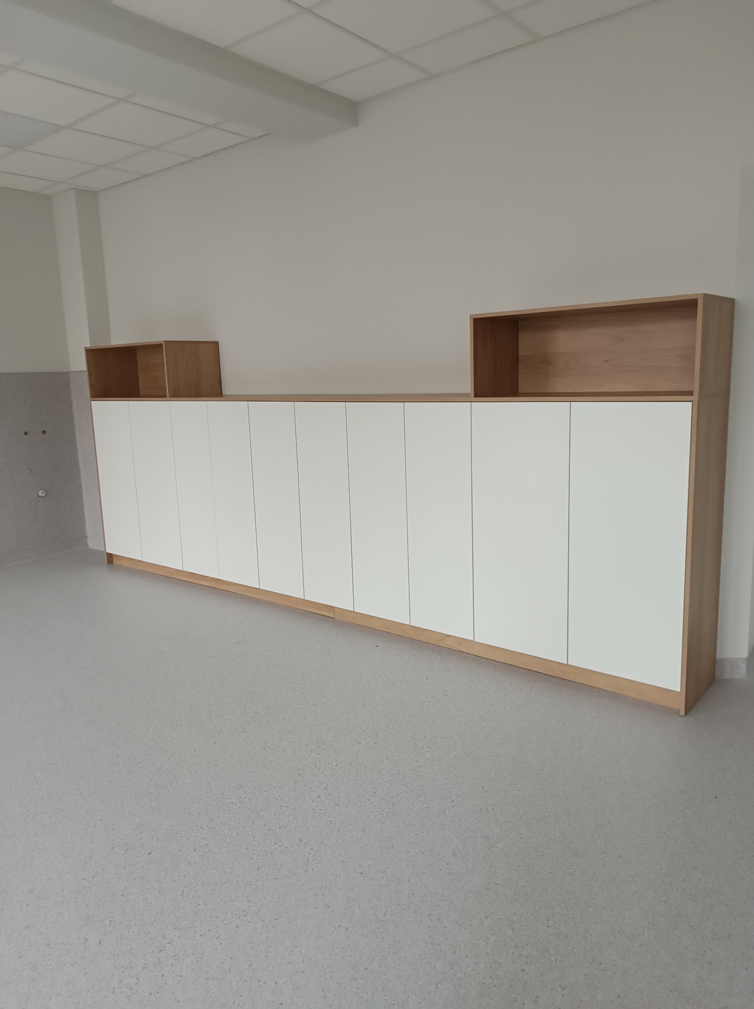 Długa szafa wnękowa z białymi frontami i drewnianymi elementami, zwieńczona otwartymi półkami, ustawiona w jasnym pomieszczeniu z szarą podłogą.