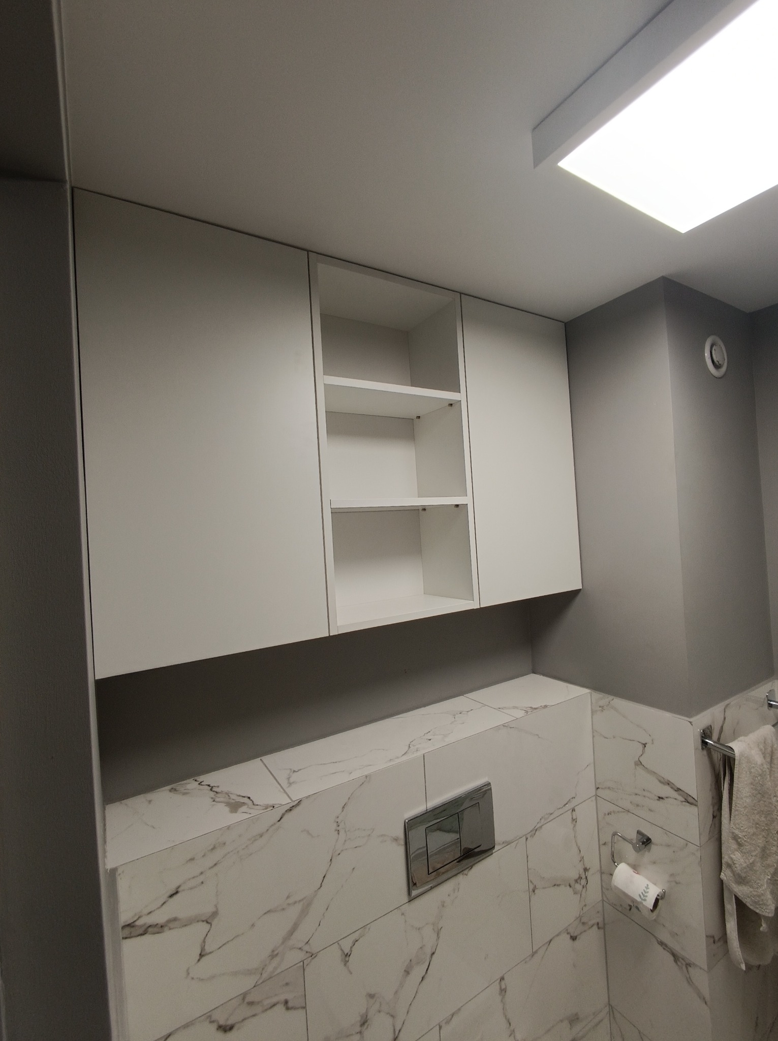 Białe szafki wiszące nad toaletą z marmurowymi płytkami, widoczne półki w jednej z szafek, minimalistyczny design.