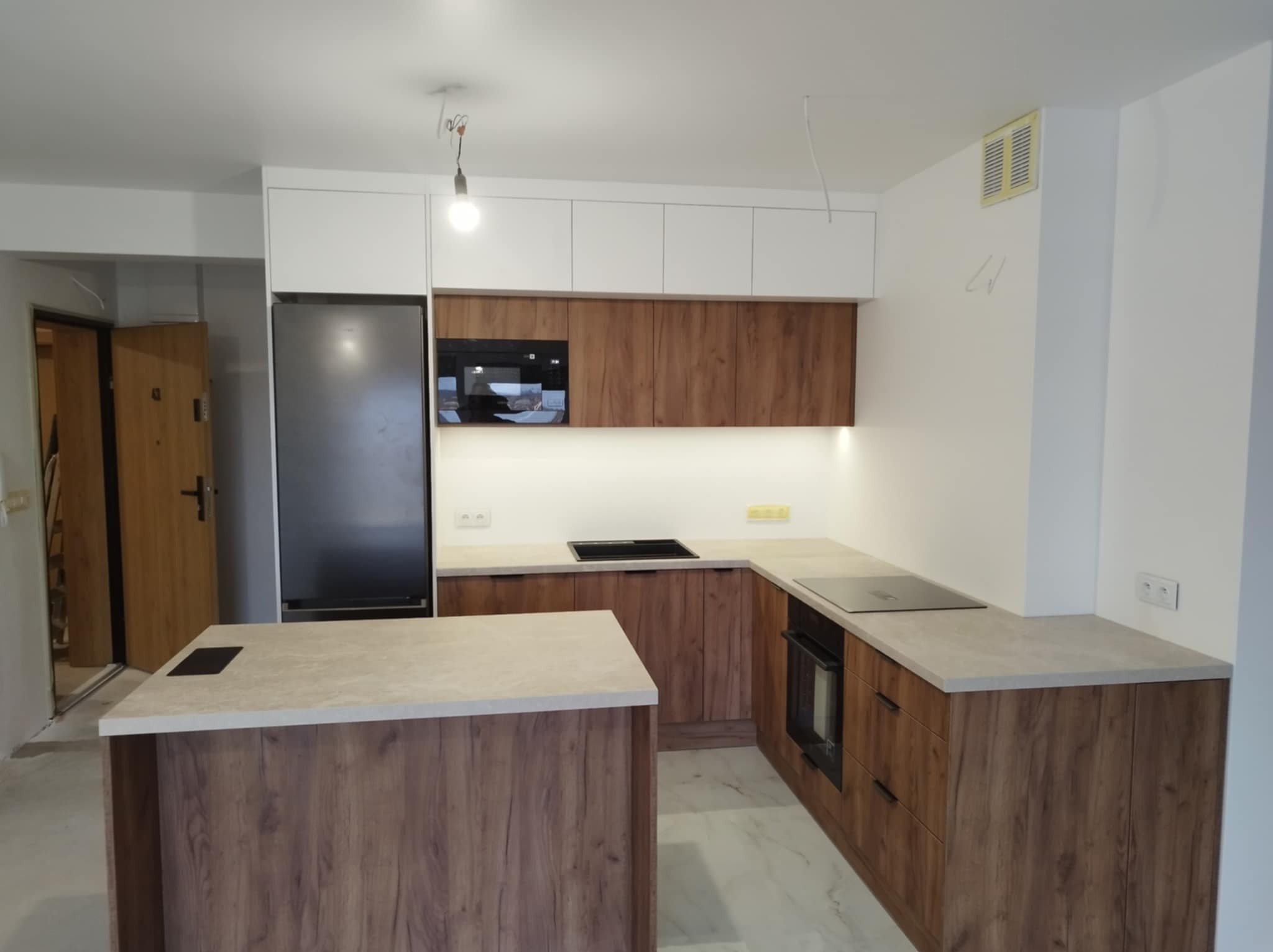 Nowoczesna kuchnia z wyspą, fronty w odcieniach drewna, białe szafki górne, czarny sprzęt AGD, marmurowe wykończenie podłogi, lampa wisząca z żarówką Edison, widoczne niedokończone prace...