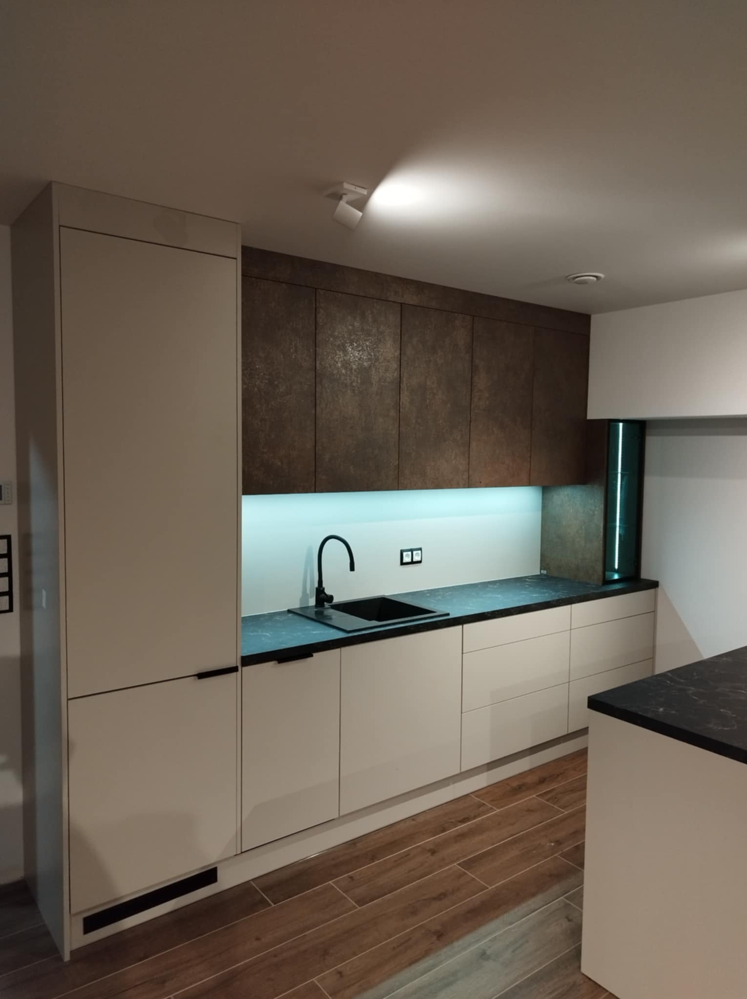 Nowoczesna kuchnia z białymi frontami szafek dolnych i brązowymi górnymi, czarnym blatem i armaturą, podświetleniem LED oraz drewnopodobną podłogą.