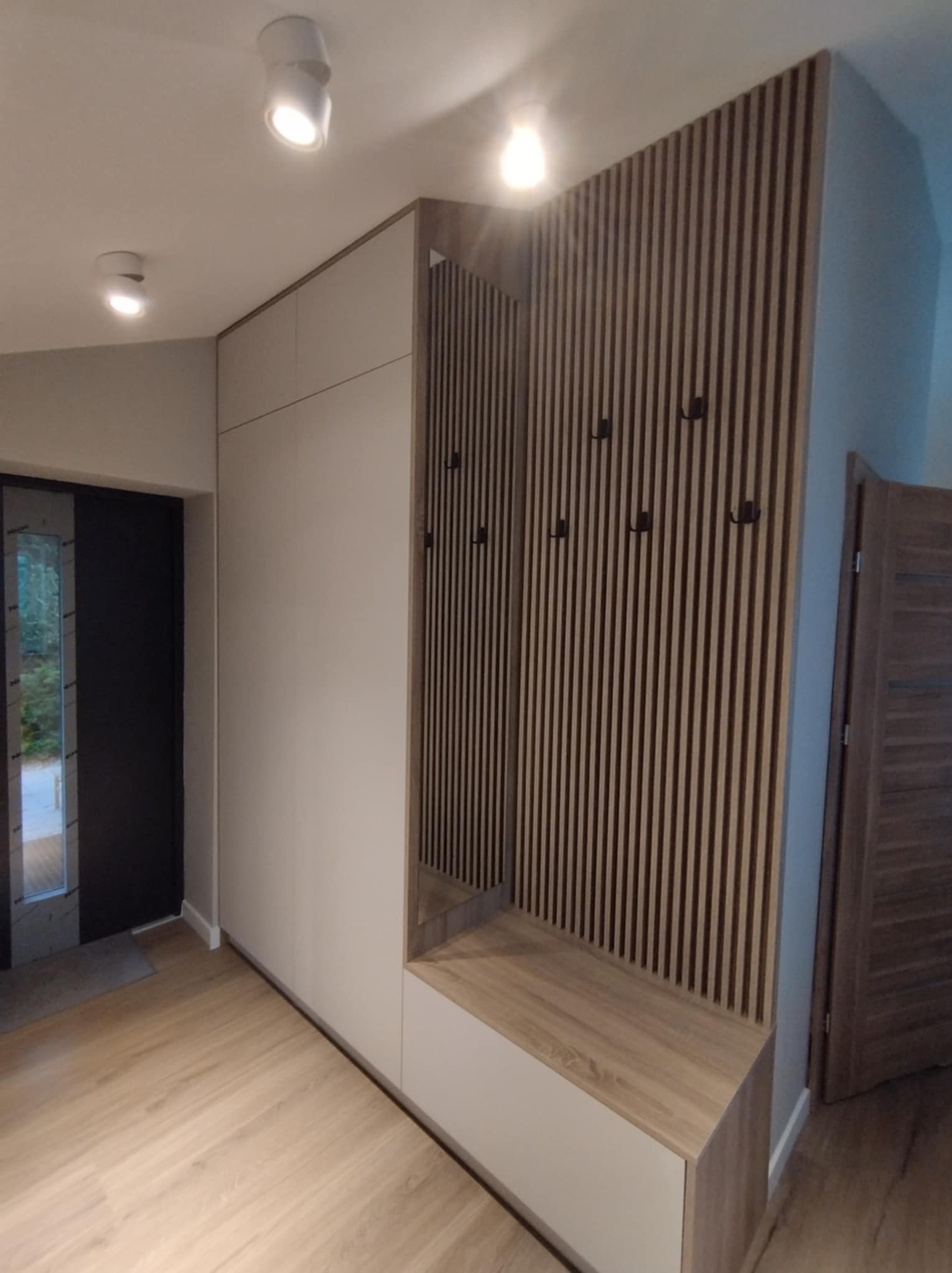 Nowoczesna szafa wnękowa z siedziskiem i panelem ściennym z wieszakami w przedpokoju w Bierutowie. Minimalistyczny design, jasne kolory, drewnopodobne wykończenie.