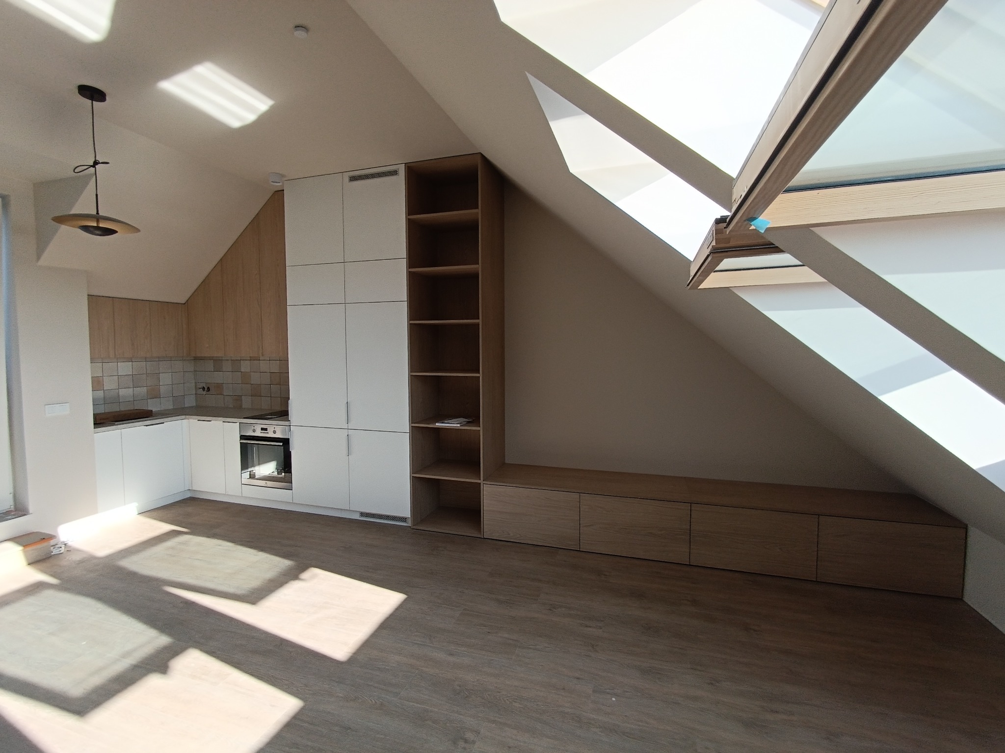 Jasna kuchnia na poddaszu ze skośnym oknem dachowym, białe szafki, drewniane akcenty, otwarte półki i zabudowa w skosie.