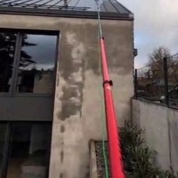 Sawielux - Mycie elewacji domu nowoczesną lancą teleskopową z zielonym wężem, widoczne ślady po czyszczeniu na szarej ścianie. Ujęcie z dołu, pochmurne niebo.