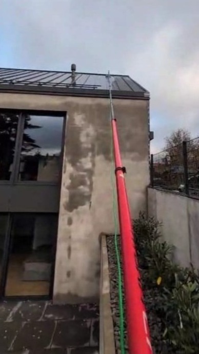 Mycie elewacji domu nowoczesną lancą teleskopową z zielonym wężem, widoczne ślady po czyszczeniu na szarej ścianie. Ujęcie z dołu, pochmurne niebo.