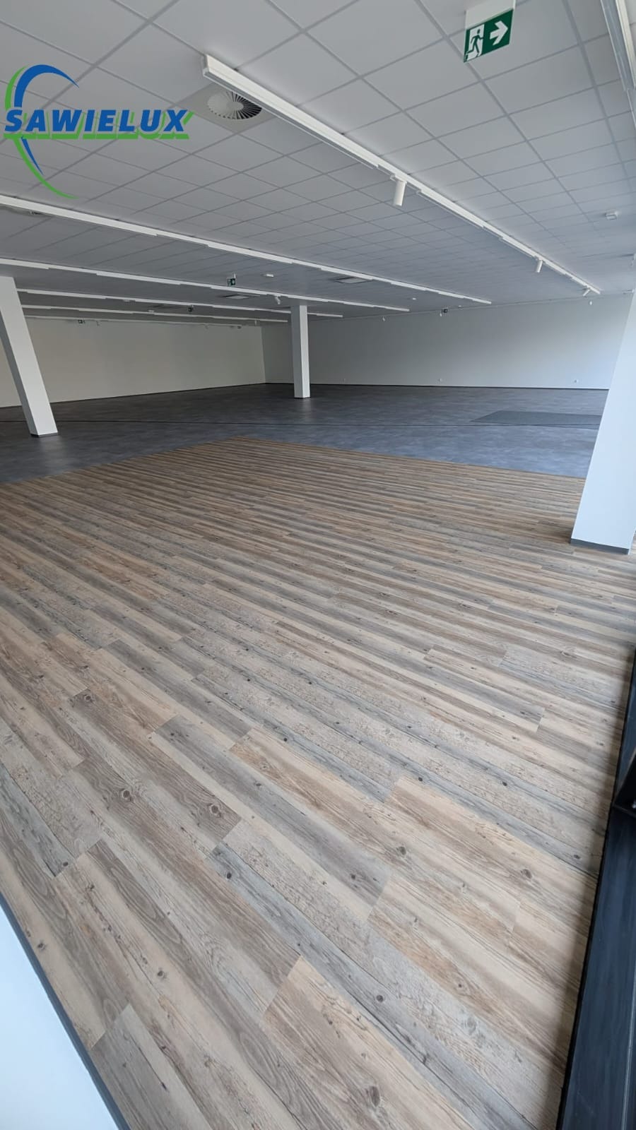 Wnętrze przestronnego, jasnego pomieszczenia z panelami podłogowymi imitującymi drewno i szarą posadzką. Widoczne białe kolumny i sufit z oświetleniem.