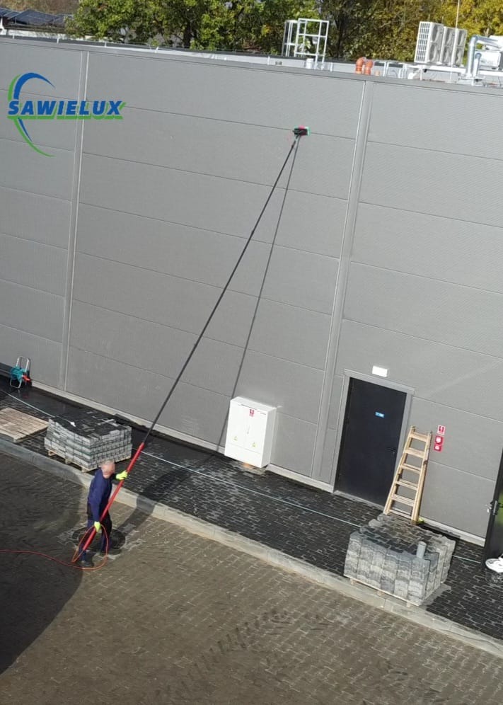 Pracownik w trakcie mycia elewacji przemysłowej za pomocą teleskopowej lancy z dyszą. Widoczna drabina, kostka brukowa i budynek z szarej blachy trapezowej.