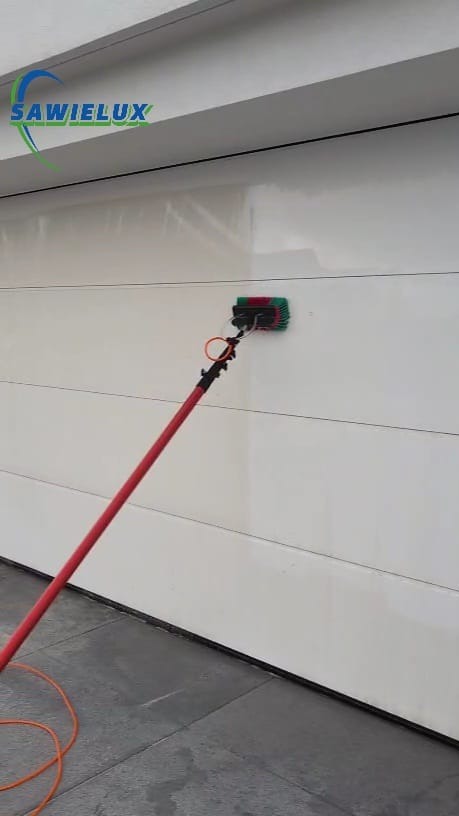 Mycie białej, poziomej bramy garażowej za pomocą szczotki na teleskopowym kiju z doprowadzeniem wody. Widoczny fragment logo firmy.