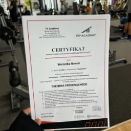 Certyfikat ukończenia kursu trenera personalnego Fit Academy, trzymany w dłoni na tle siłowni z maszynami do ćwiczeń i ćwiczącymi osobami.