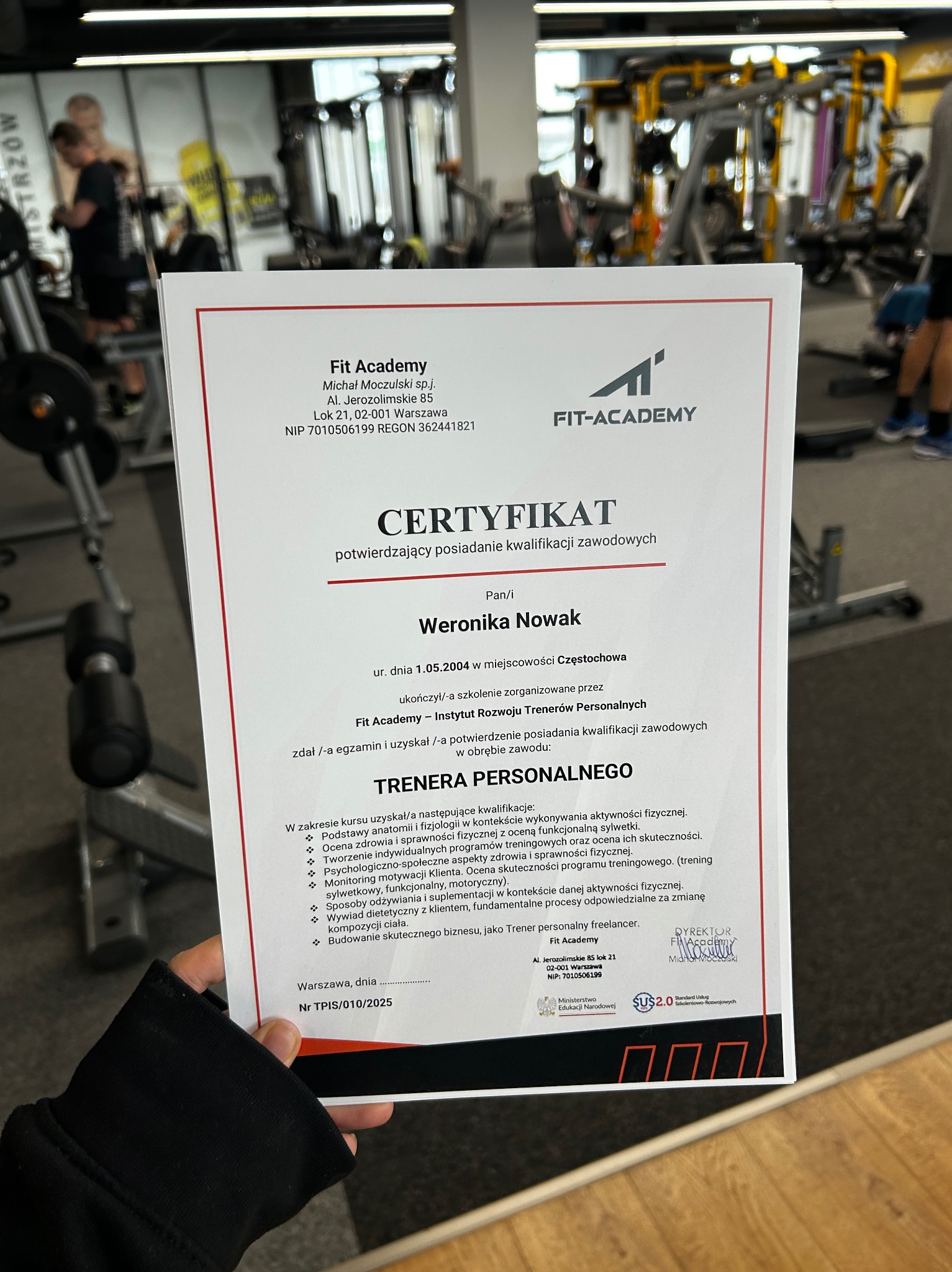 Certyfikat ukończenia kursu trenera personalnego Fit Academy, trzymany w dłoni na tle siłowni z maszynami do ćwiczeń i ćwiczącymi osobami.