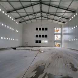 OneXpol - Wnętrze nowo wybudowanej hali z widoczną konstrukcją stalową dachu, ścianami z paneli warstwowych i betonową posadzką z plamami wody. Podnośnik koszowy widoczny w otworze okiennym.