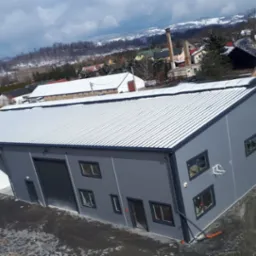 Nowoczesna hala przemysłowa z szarej płyty warstwowej, widok z góry, otwarte bramy wjazdowe, w tle zimowy krajobraz z ośnieżonymi wzgórzami.