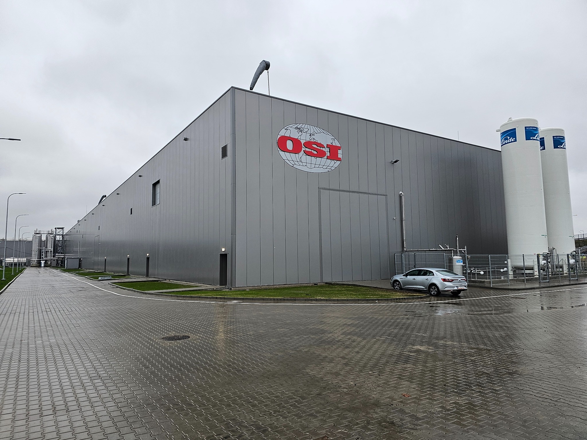 Szara hala przemysłowa z logo OSI na elewacji, widoczne zbiorniki Linde oraz zaparkowany samochód, na mokrym brukowanym placu.