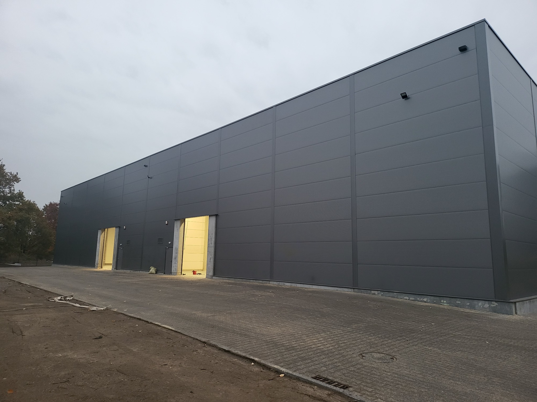 Szara hala przemysłowa z płyt warstwowych z dwoma otwartymi bramami wjazdowymi, widoczny fragment terenu przed budynkiem i pochmurne niebo.