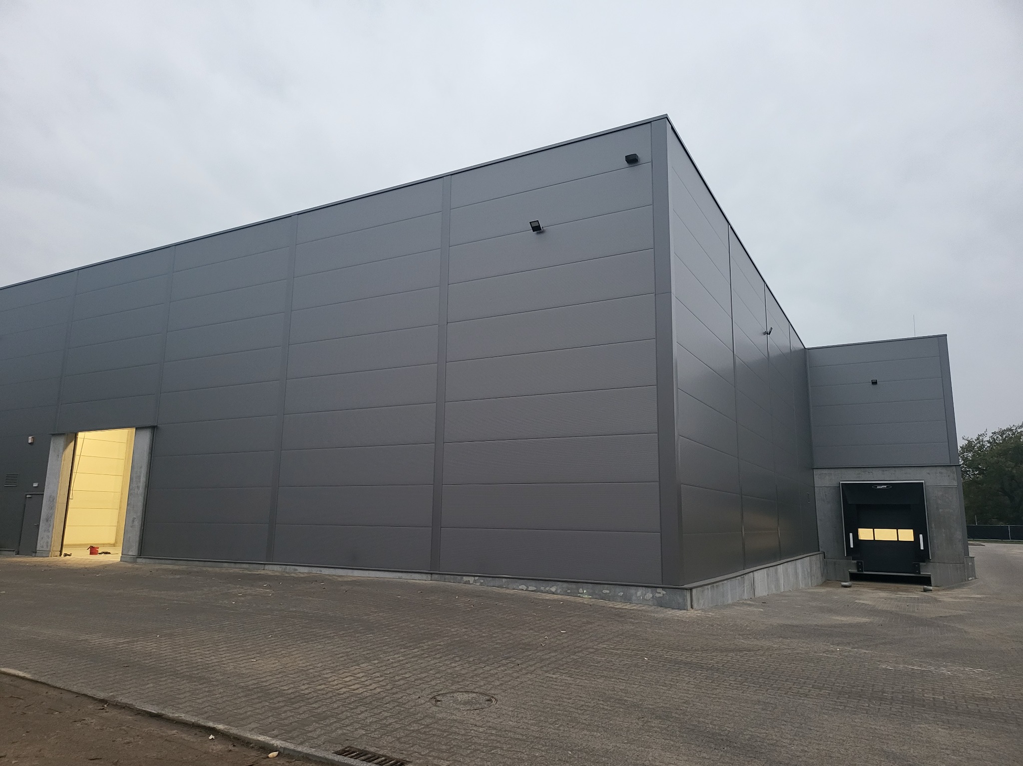 Nowoczesna hala przemysłowa z szarych płyt warstwowych, widoczne otwarte wrota i brama załadunkowa, pochmurne niebo w tle.