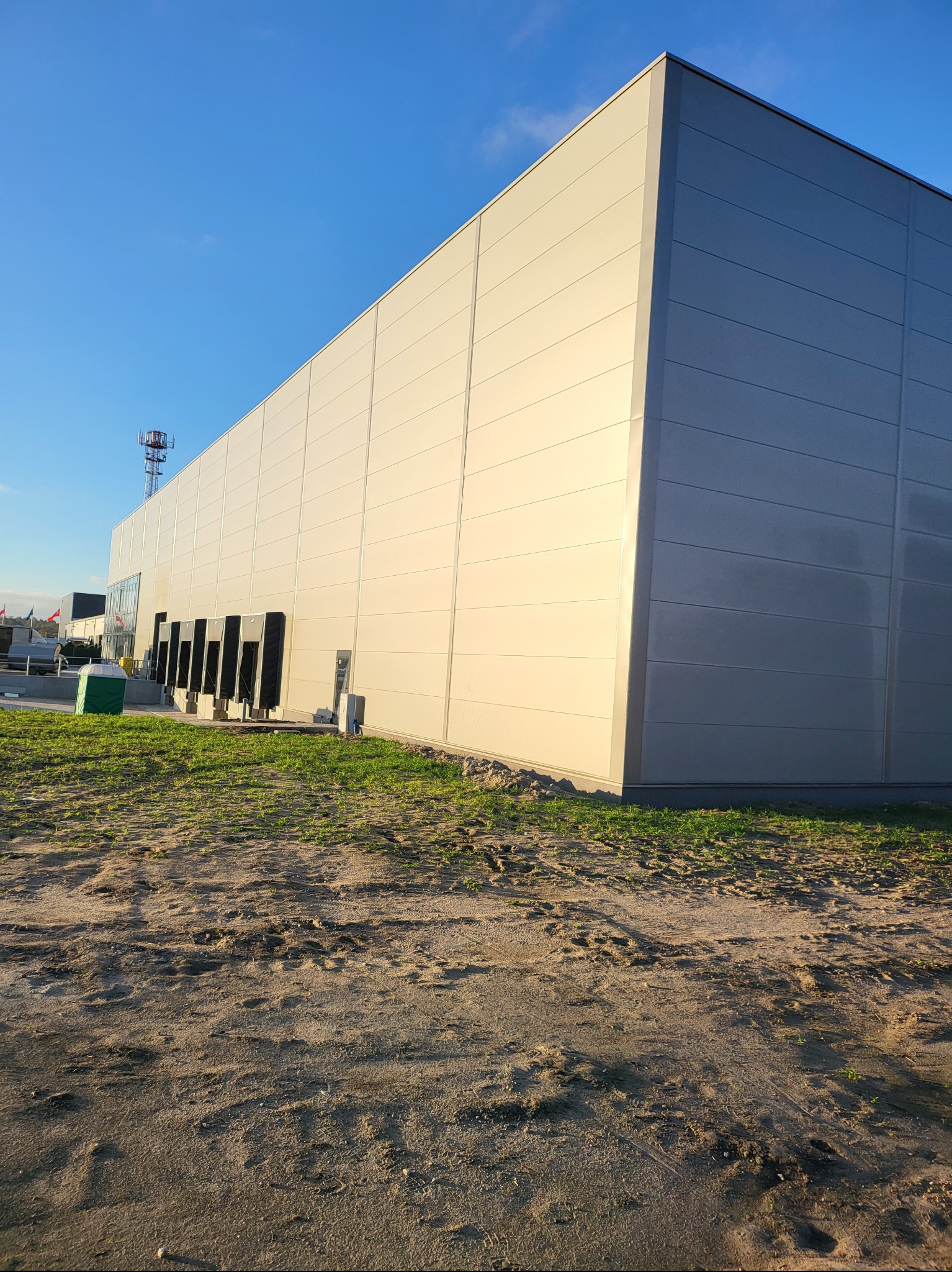 Kątowe ujęcie hali przemysłowej z jasnych płyt warstwowych z widocznymi dokami załadunkowymi, na pierwszym planie teren z elementami trawy, w tle maszt telekomunikacyjny i fragmenty innych budynków.