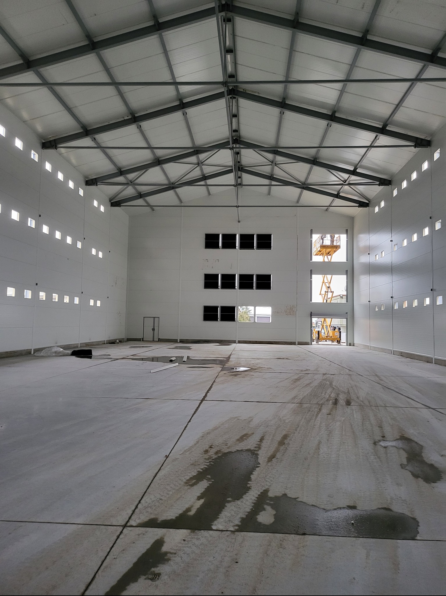 Wnętrze nowo wybudowanej hali z widoczną konstrukcją stalową dachu, ścianami z paneli warstwowych i betonową posadzką z plamami wody. Podnośnik koszowy widoczny w otworze okiennym.