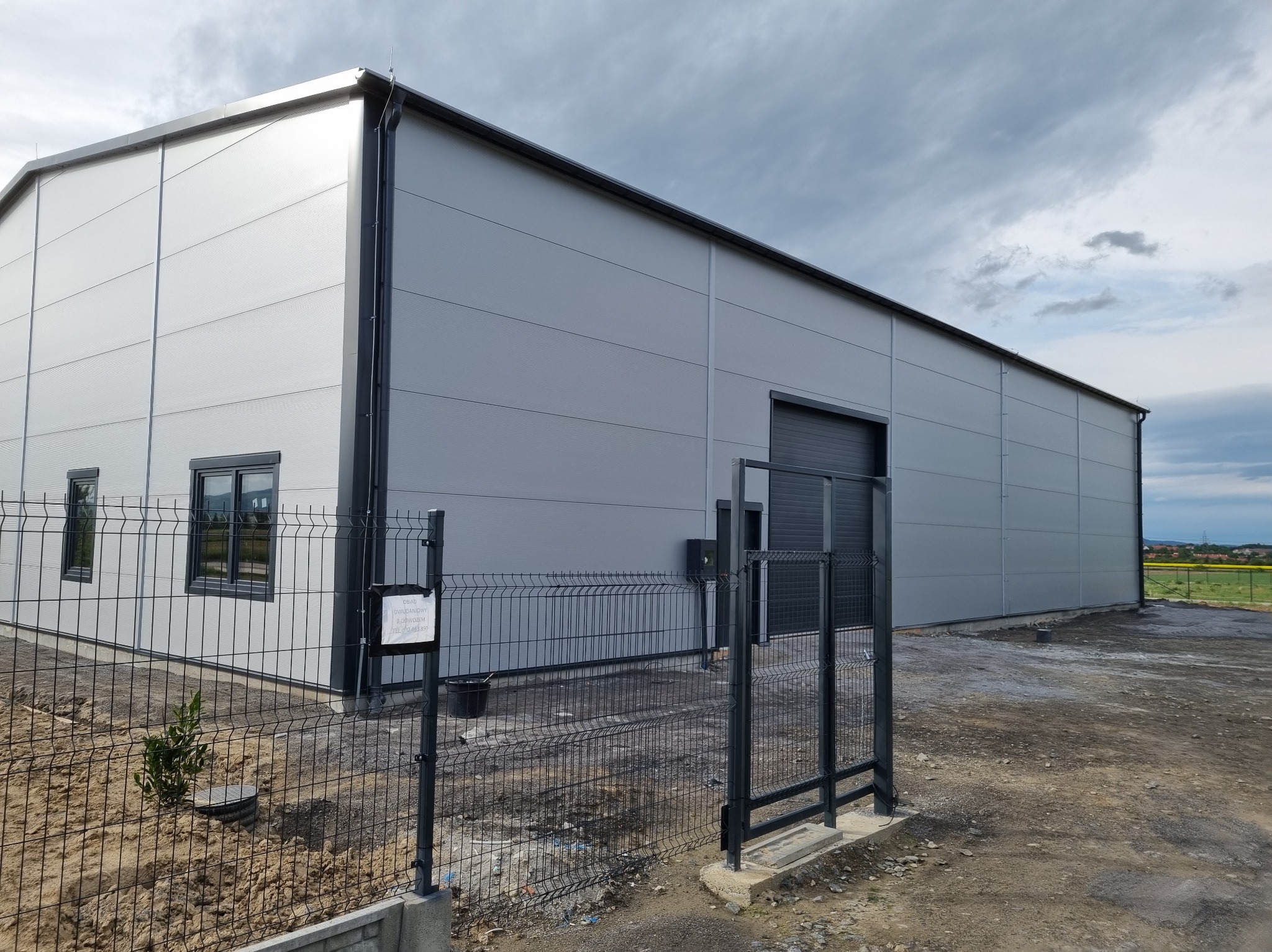 Nowoczesna hala przemysłowa z szarych płyt warstwowych, widoczna boczna ściana z oknami i brama wjazdowa, ogrodzona siatką z widocznym ogłoszeniem na słupku.