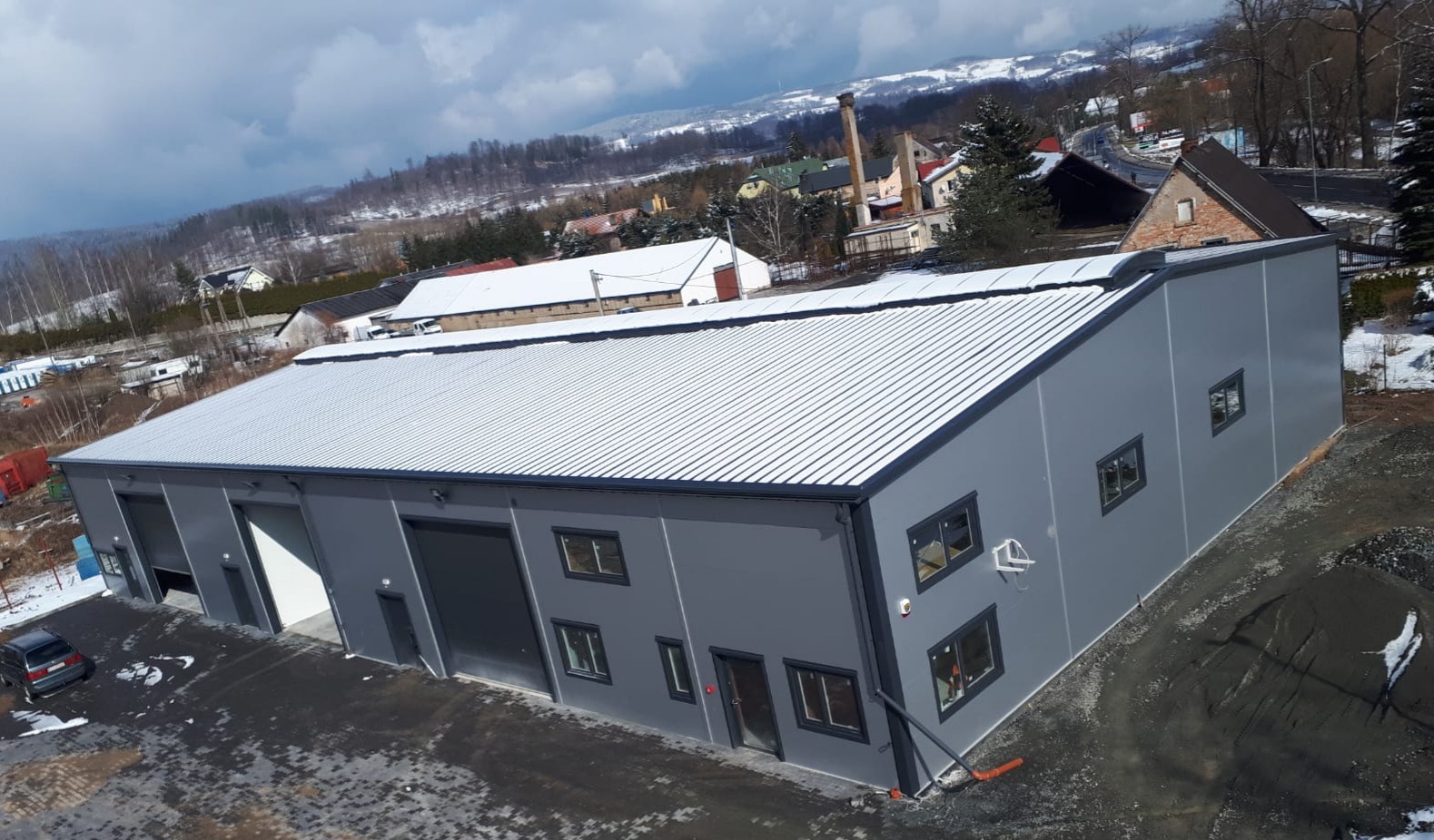 Nowoczesna hala przemysłowa z szarej płyty warstwowej, widok z góry, otwarte bramy wjazdowe, w tle zimowy krajobraz z ośnieżonymi wzgórzami.