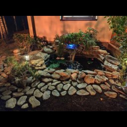 Duodym - Ogród nocą z oświetlonym oczkiem wodnym, kamienną kaskadą i ścieżką z nieregularnych płyt, widoczne rośliny ozdobne i lampa ogrodowa.