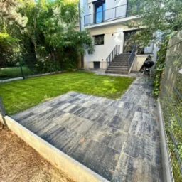 Ułożona kostka brukowa na podw&oacute;rku z widokiem na świeży trawnik i budynek z balkonem, całość otoczona płotem z siatki.