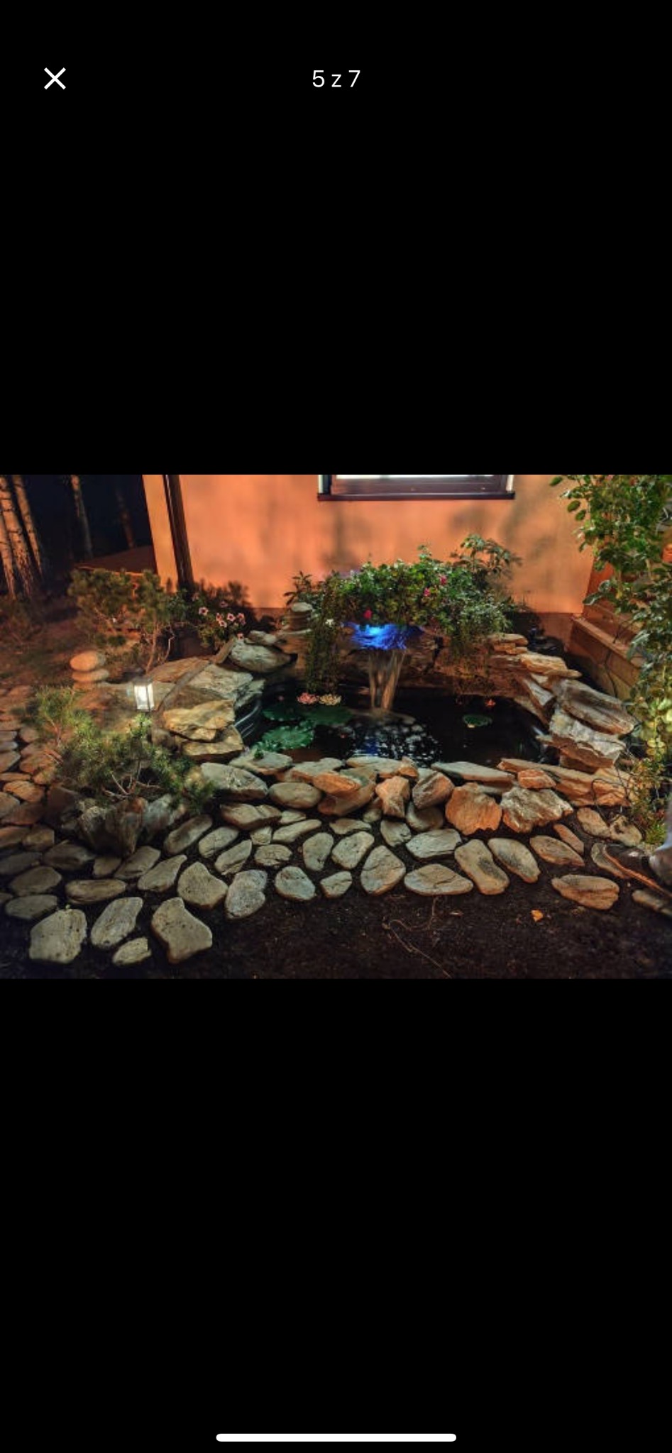 Ogród nocą z oświetlonym oczkiem wodnym, kamienną kaskadą i ścieżką z nieregularnych płyt, widoczne rośliny ozdobne i lampa ogrodowa.