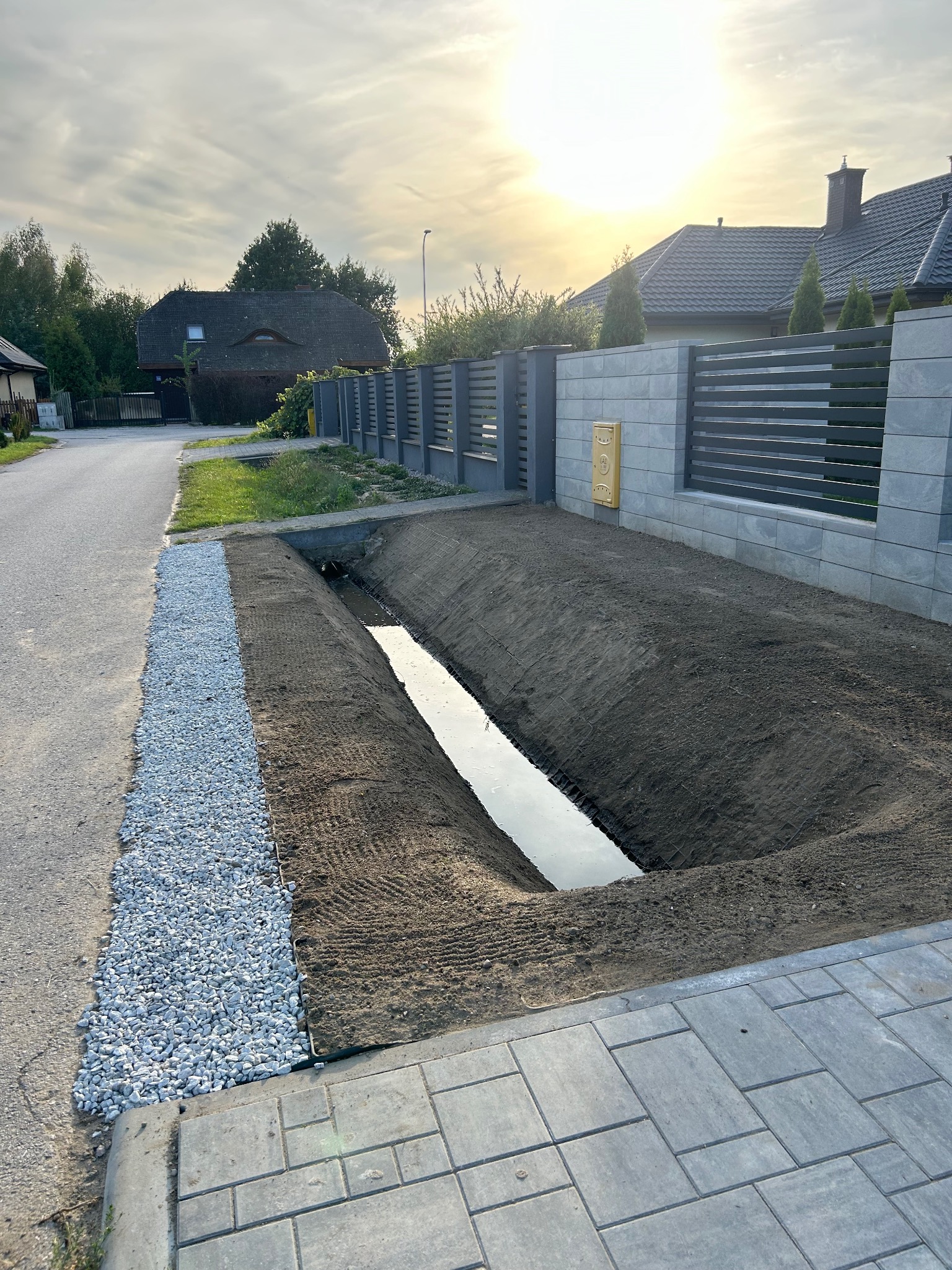 Wykonany wykop pod drenaż wzdłuż ogrodzenia z ułożonym obrzeżem z kamienia, w tle zabudowa jednorodzinna.