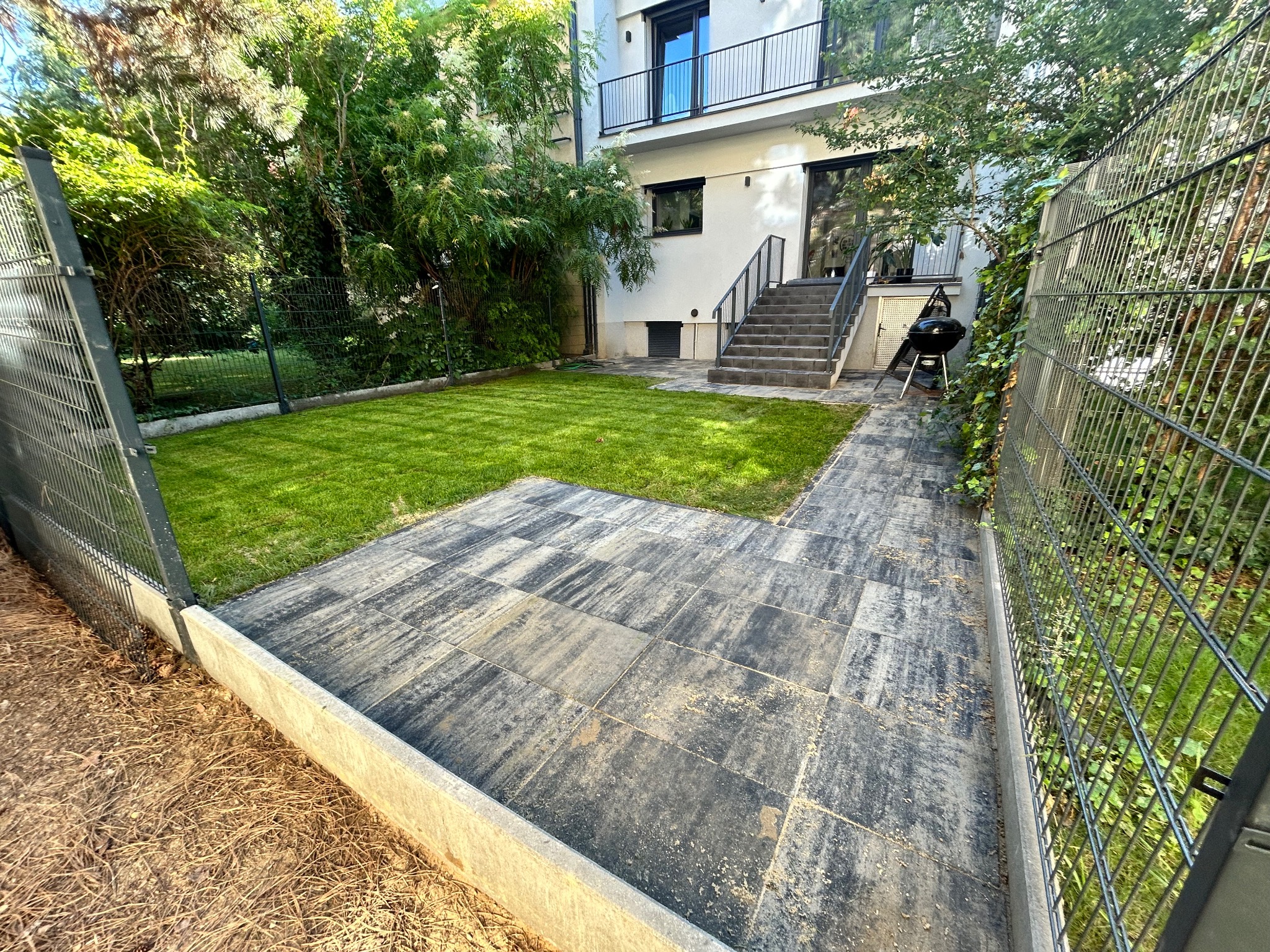 Ułożona kostka brukowa na podwórku z widokiem na świeży trawnik i budynek z balkonem, całość otoczona płotem z siatki.