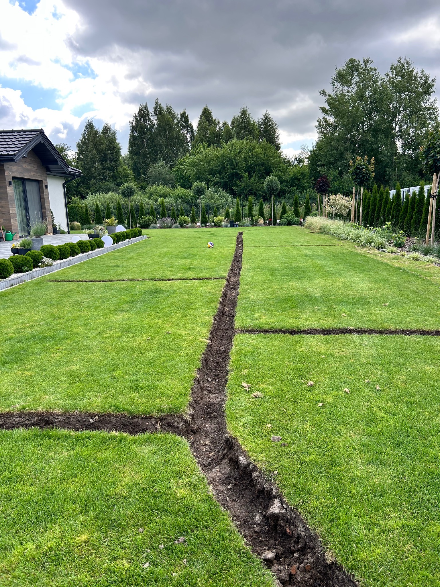 Wykonane rowy w trawniku pod przyszłą instalację nawadniającą. Widoczne równe linie cięcia darni, regularny kształt trawnika i elementy architektury ogrodowej w tle.