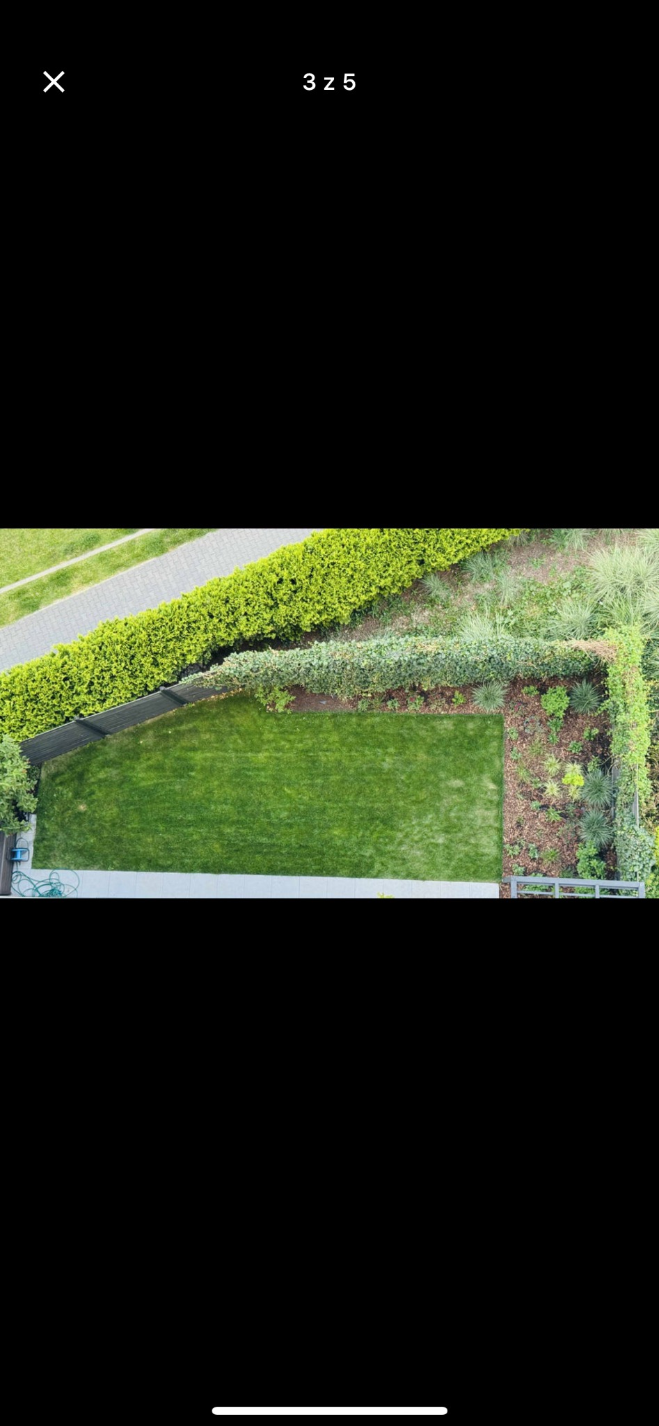 Widok z góry na zielony trawnik otoczony żywopłotem i rabatami, z elementami architektury ogrodowej, ukazujący zadbany i uporządkowany krajobraz.