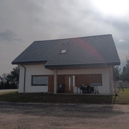 Darfiko - Nowoczesny dom jednorodzinny z ciemnoszarą dachówką, białymi ścianami i drewnianą okładziną elewacyjną, widoczny z perspektywy podjazdu w pochmurny dzień.