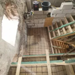 Budowa fundamentów: zbrojenie siatką stalową i szalunek drewniany w wykopie o nierównych ścianach z cegły i betonu, widoczne narzędzia i materiały budowlane.