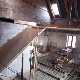 Wnętrze poddasza w trakcie adaptacji: widoczne białe bloczki, drewniana konstrukcja dachu z oknem połaciowym, odsłonięte ceglane ściany, narzędzia i materiały budowlane na podłodze.