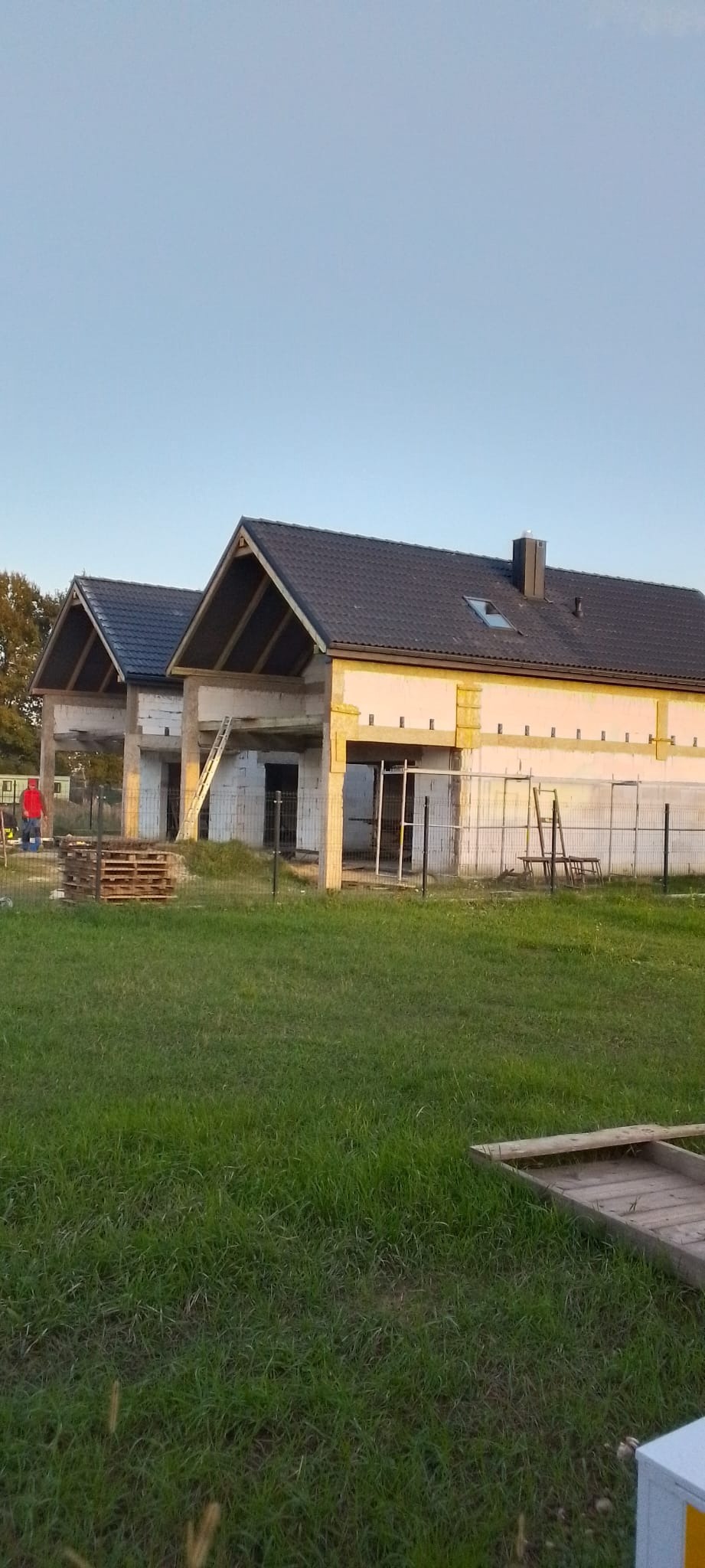 Dwa budynki w trakcie budowy w Marcinowicach, z czarnymi dachami i ocieplonymi ścianami, widoczne rusztowania i materiały budowlane na trawniku.