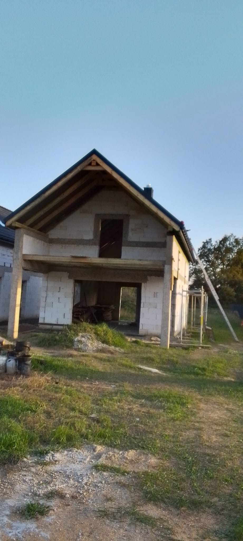 Wznoszony dom z białych bloczków, z częściowo ukończonym dachem i widocznymi drewnianymi elementami konstrukcyjnymi, z drabiną opartą o ścianę, na tle zielonego trawnika i błękitnego nieba.