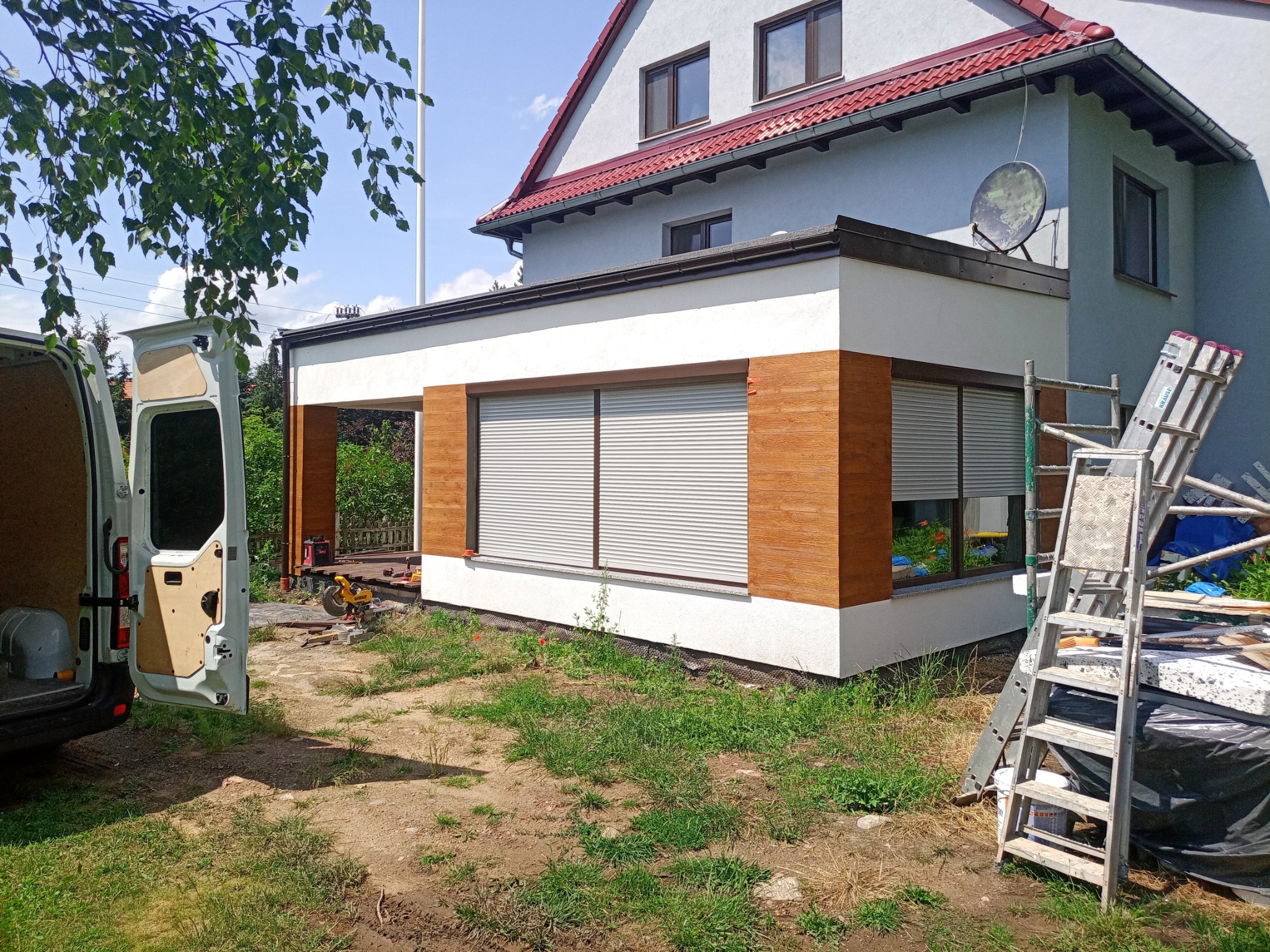 Rozbudowa domu jednorodzinnego z drewnianą elewacją, widoczne rolety zewnętrzne, drabina oparta o ścianę, otwarte drzwi samochodu dostawczego z narzędziami w tle.