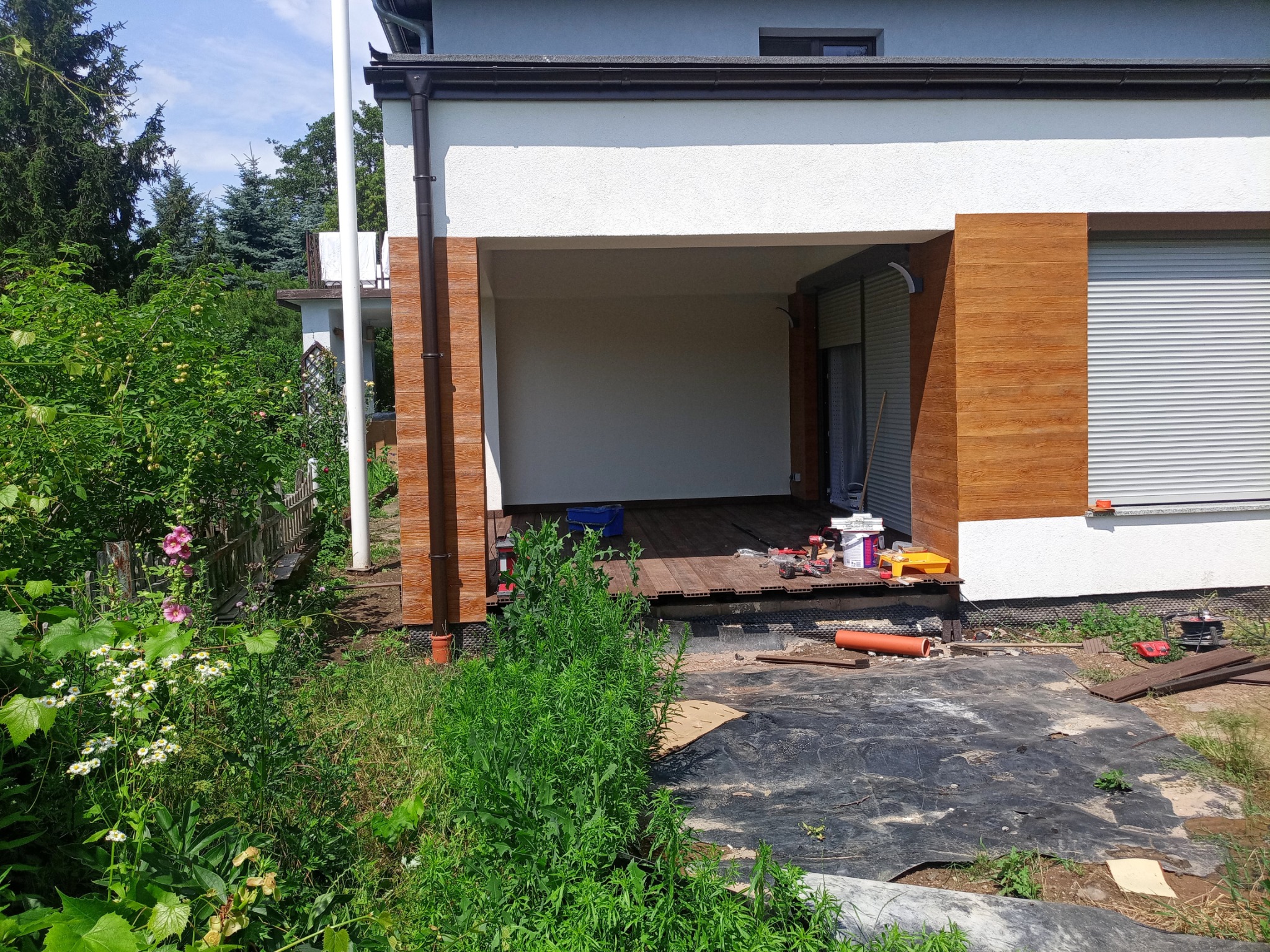 Budowa zadaszonego tarasu przy domu: widoczne deski tarasowe, narzędzia, folia ochronna na gruncie i fragment ogrodu.