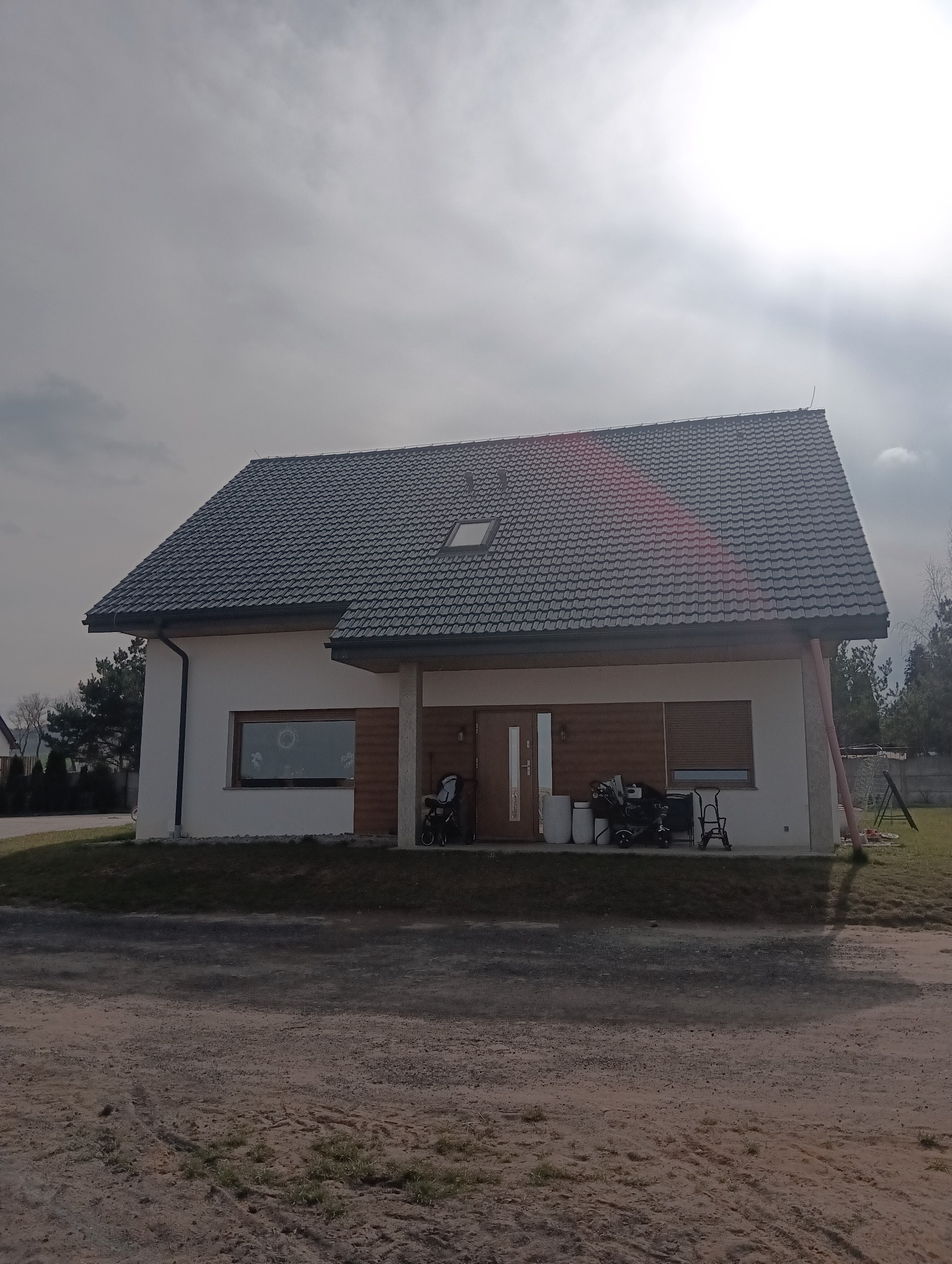 Nowoczesny dom jednorodzinny z ciemnoszarą dachówką, białymi ścianami i drewnianą okładziną elewacyjną, widoczny z perspektywy podjazdu w pochmurny dzień.