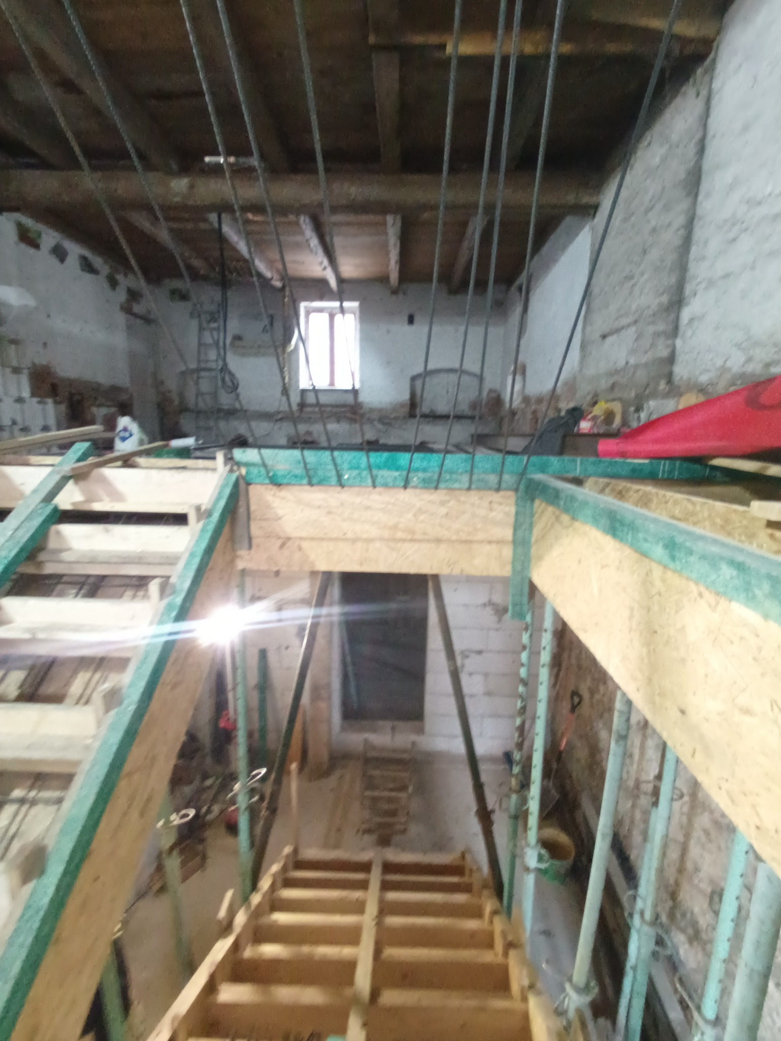 Wnętrze remontowanego budynku z drewnianą konstrukcją podtrzymującą strop, widok od dołu z drewnianymi schodami prowadzącymi do góry, w tle widoczne okno i surowe ściany z cegły oraz drewniany strop.