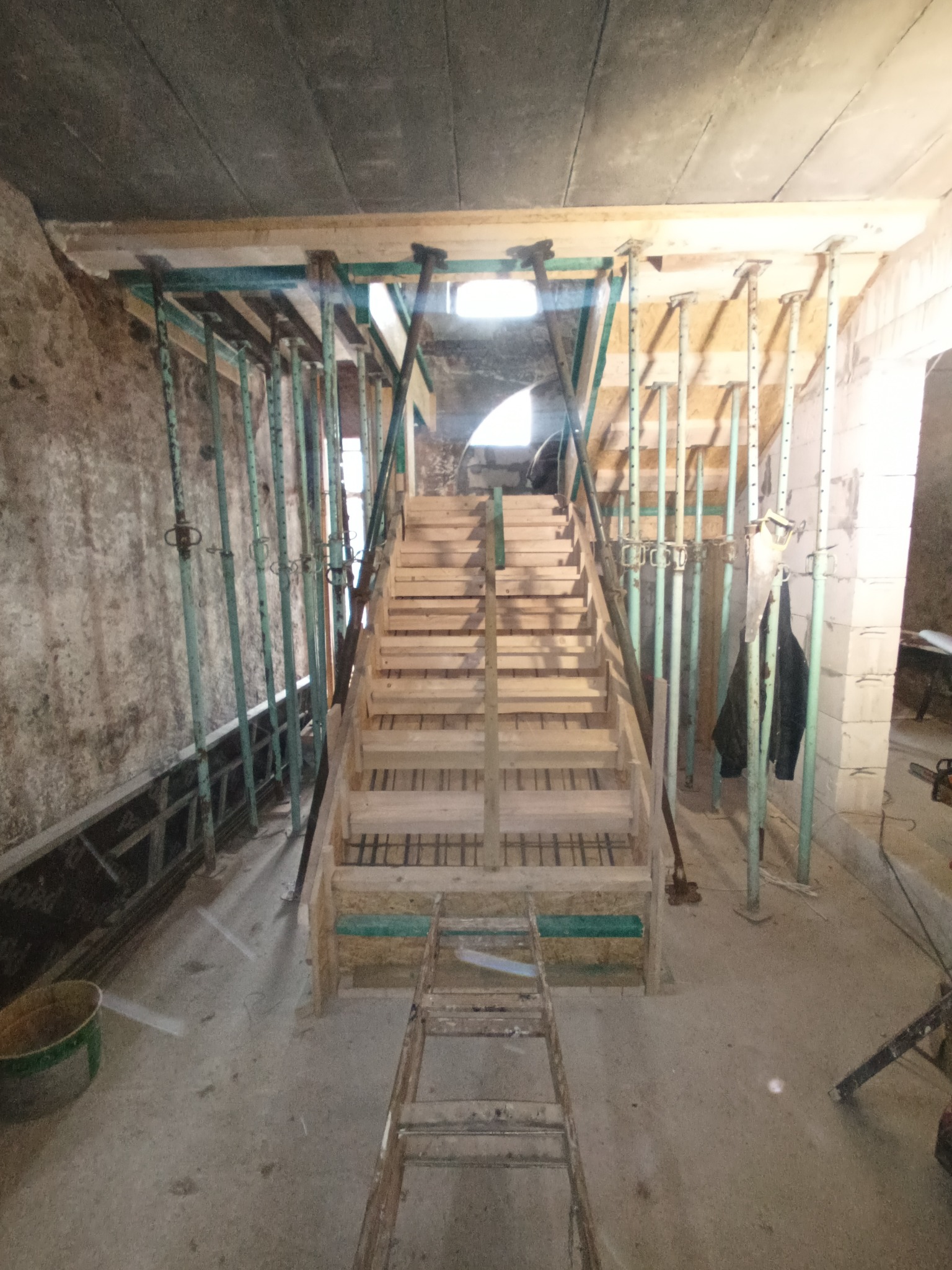 Konstrukcja szalunkowa pod schody z drewnianymi stopniami, wsparta na metalowych stemplach budowlanych, widok z dołu na surowym stanie budynku.
