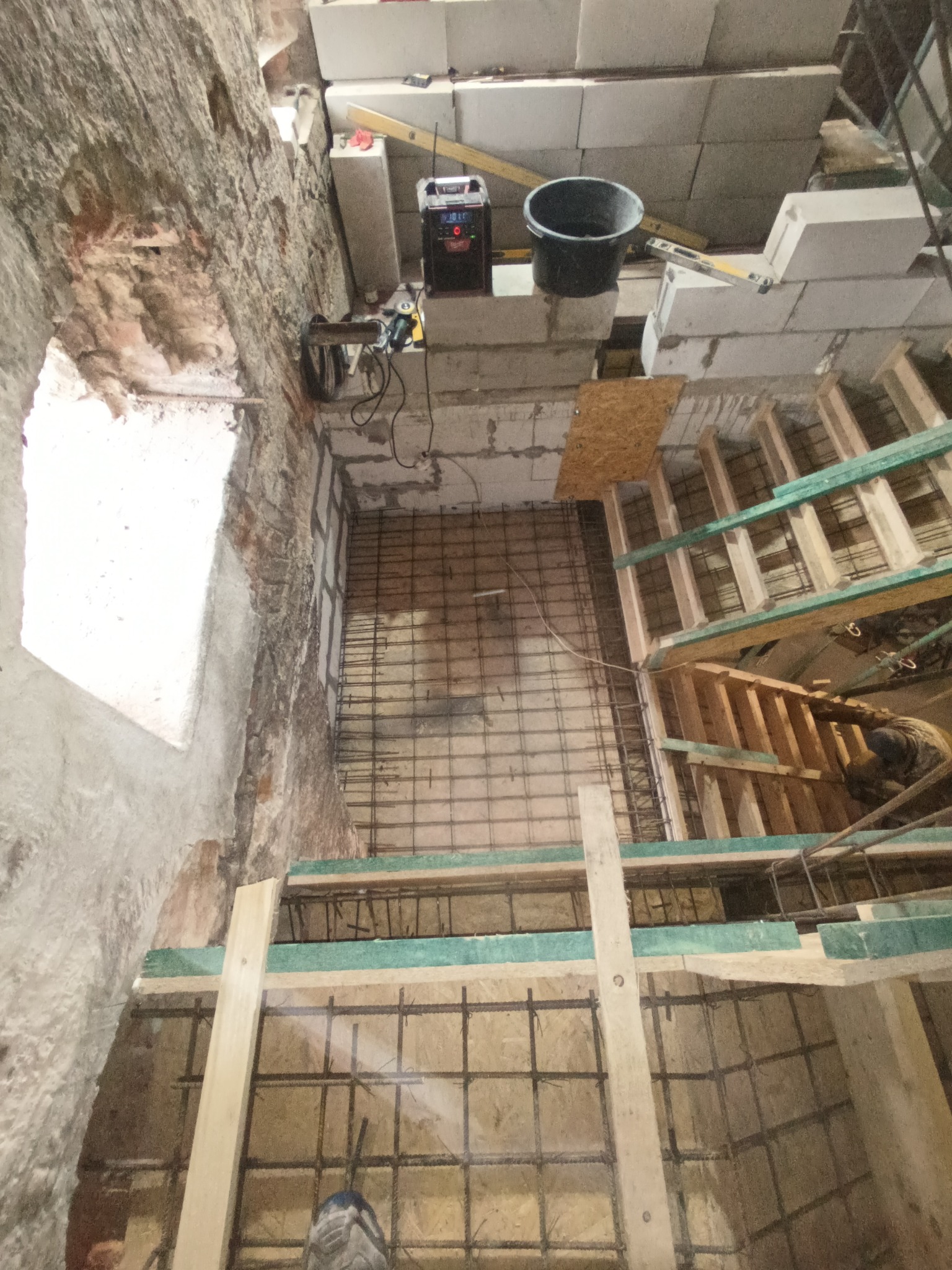 Budowa fundamentów: zbrojenie siatką stalową i szalunek drewniany w wykopie o nierównych ścianach z cegły i betonu, widoczne narzędzia i materiały budowlane.