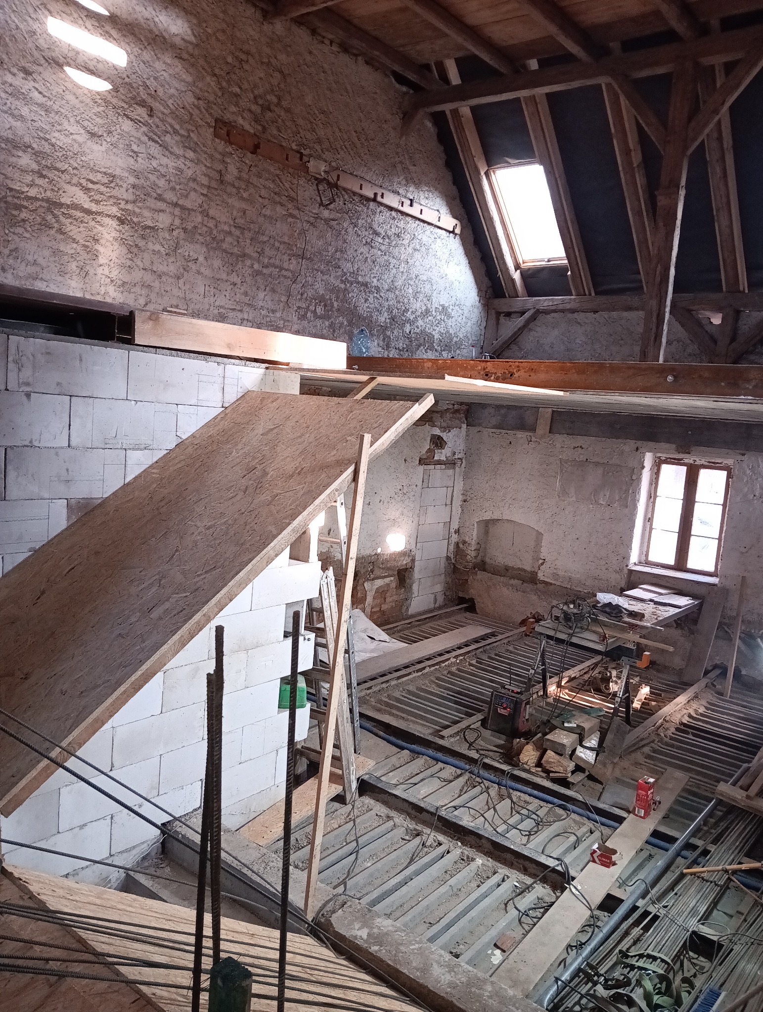 Wnętrze poddasza w trakcie adaptacji: widoczne białe bloczki, drewniana konstrukcja dachu z oknem połaciowym, odsłonięte ceglane ściany, narzędzia i materiały budowlane na podłodze.