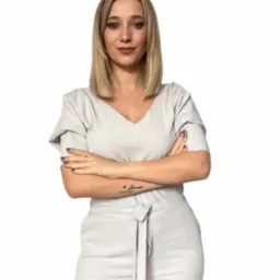 Portret kobiety z blond włosami, ubranej w jasny komplet, z tatuażem na przedramieniu, pozującej z założonymi rękami na białym tle. Profesjonalny wizerunek.