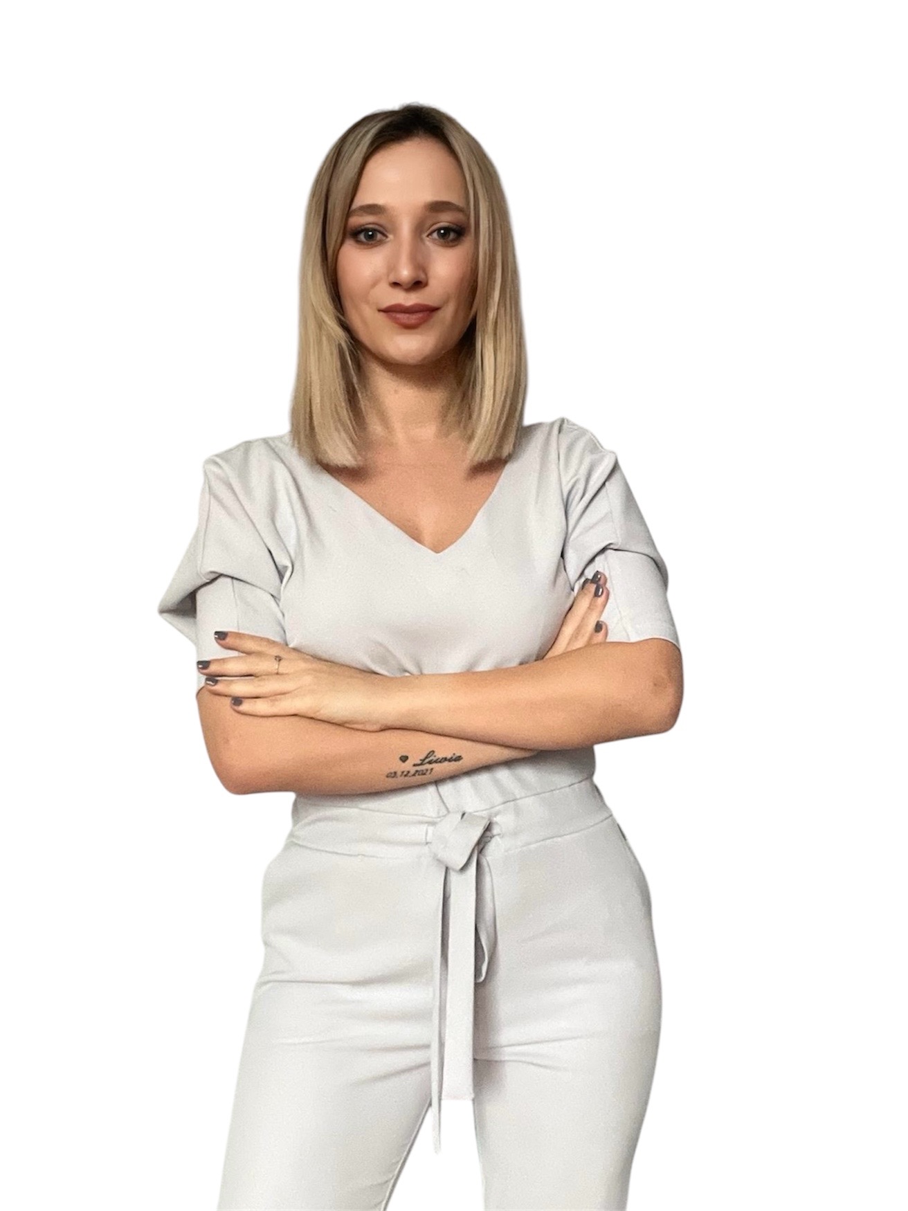 Portret kobiety z blond włosami, ubranej w jasny komplet, z tatuażem na przedramieniu, pozującej z założonymi rękami na białym tle. Profesjonalny wizerunek.