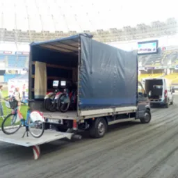 Ciężarówka z plandeką i wysuwaną rampą załadowaną rowerami miejskimi na tle stadionu sportowego, drugi bus w tle, dwóch mężczyzn rozmawia obok.