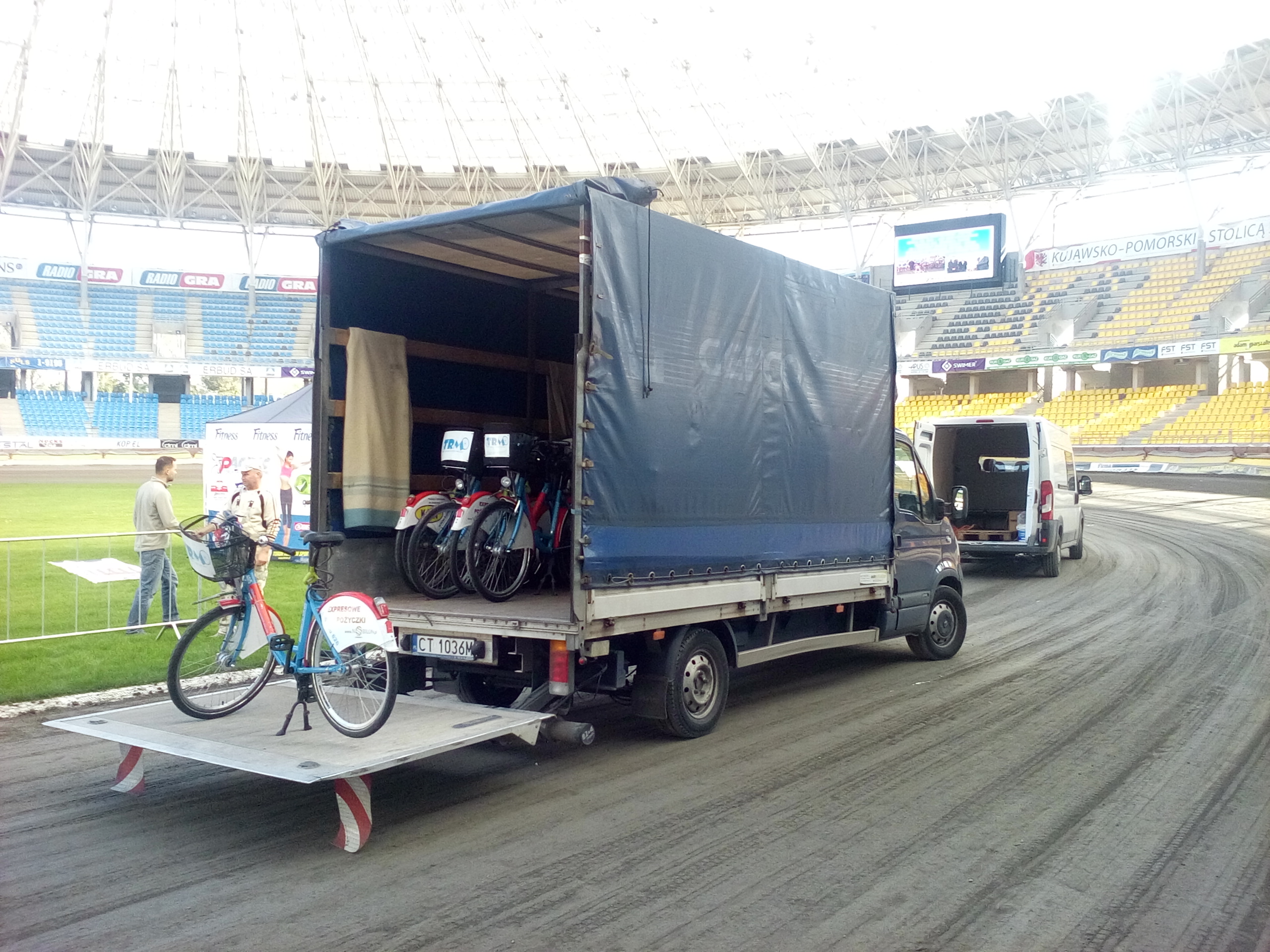 Ciężarówka z plandeką i wysuwaną rampą załadowaną rowerami miejskimi na tle stadionu sportowego, drugi bus w tle, dwóch mężczyzn rozmawia obok.